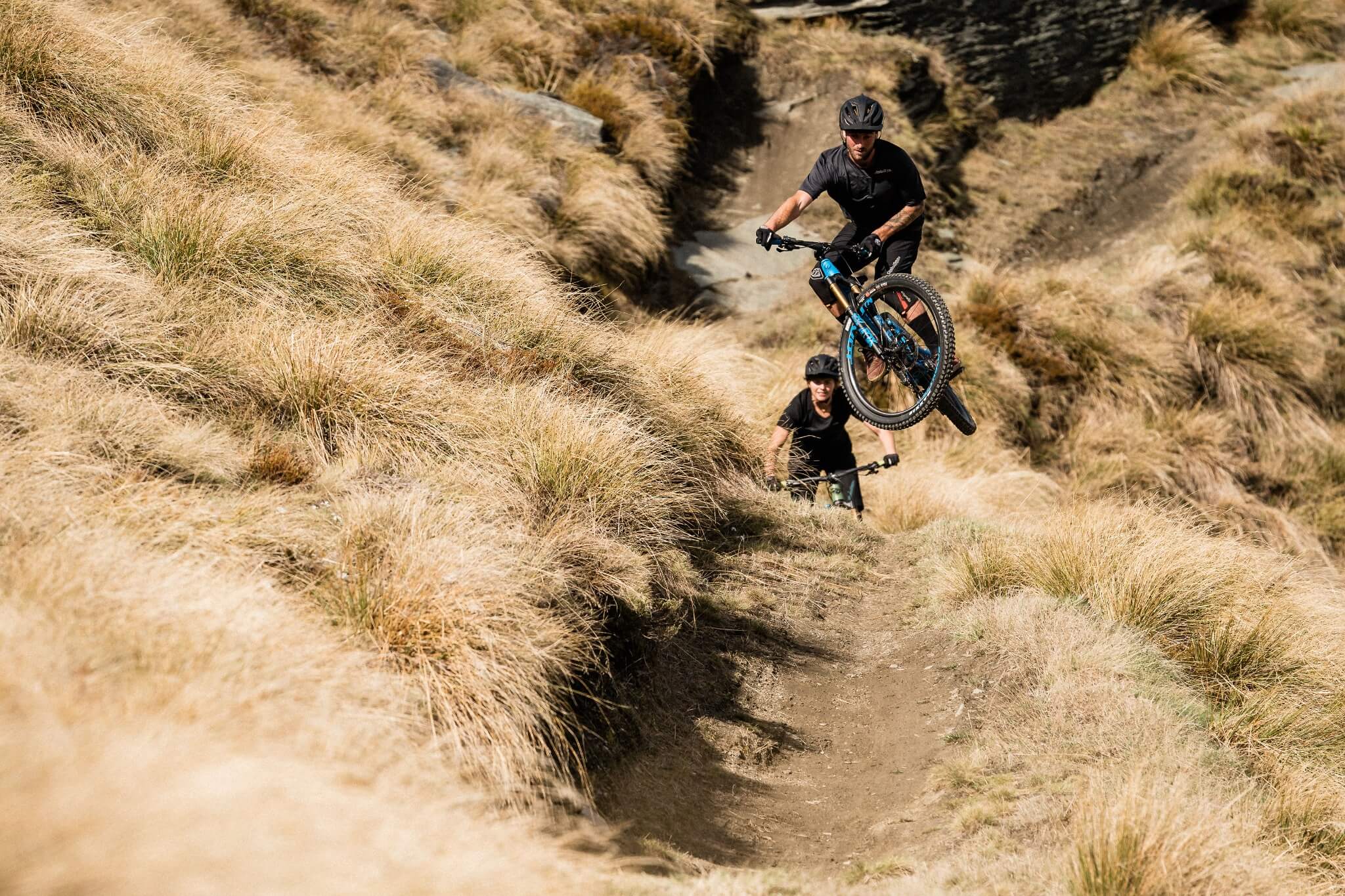 Zwei Mountainbiker fahren im Gelände; einer springt mit einem blau-schwarzen Fahrrad namens Pivot Switchblade.
