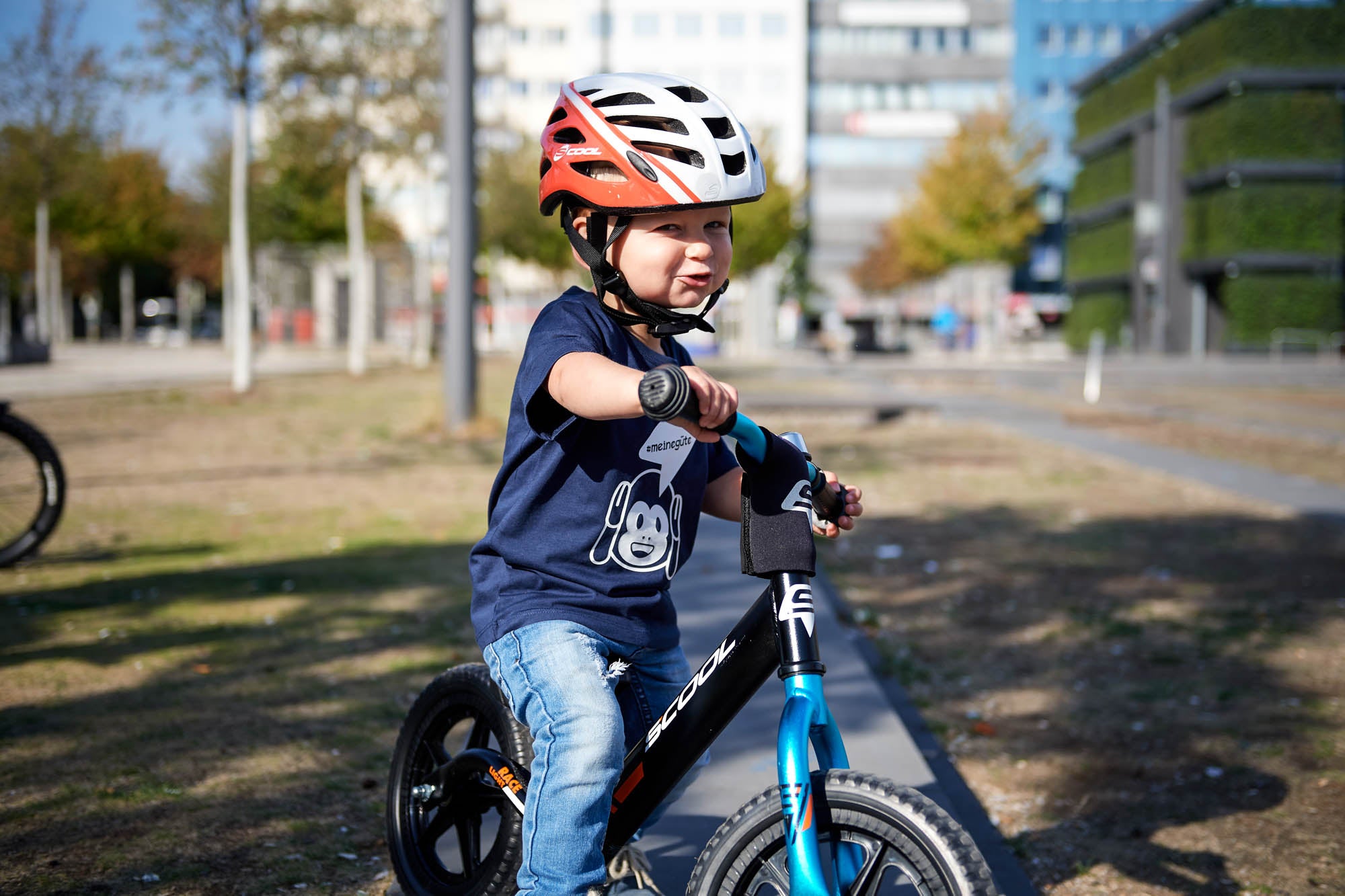 Kind auf S'cool Laufrad mit rotem Helm im Park, geeignet für junge Radfahrer.