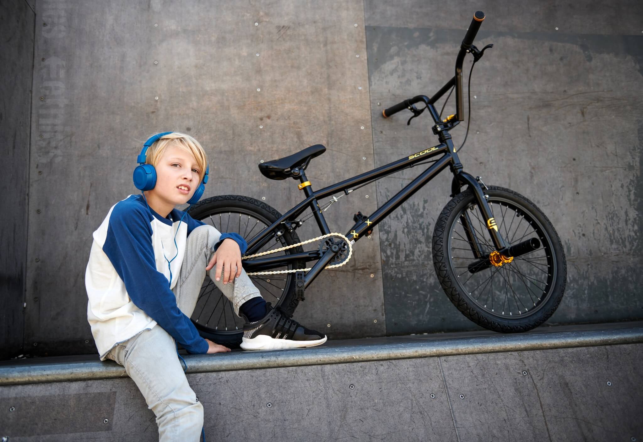 Junge mit blauen Kopfhörern sitzt neben einem schwarzen S'COOL BMX-Fahrrad auf einer Skate-Rampe.