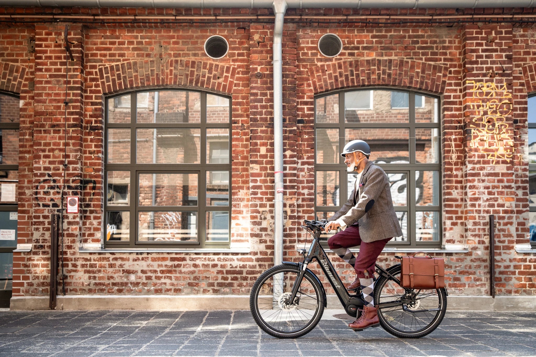 Mann auf Diamant E-Bike mit Helm, fährt vor Backsteinfassade. Herrenmode, Ledertasche, urbaner Hintergrund.