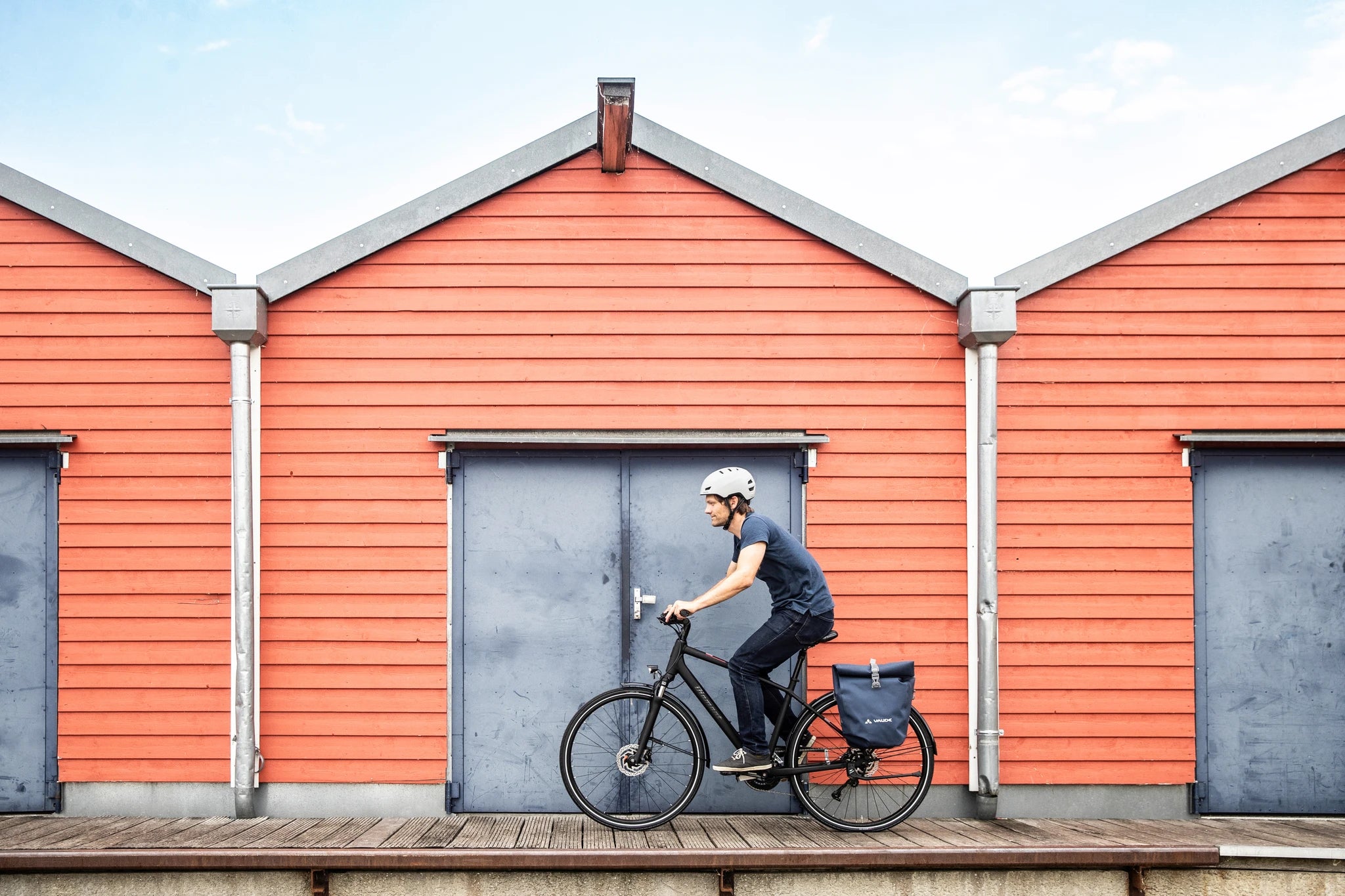 Person auf einem schwarzen Citybike mit Helm vor roten Gebäuden. Blaue Fahrradtasche am Gepäckträger befestigt.
