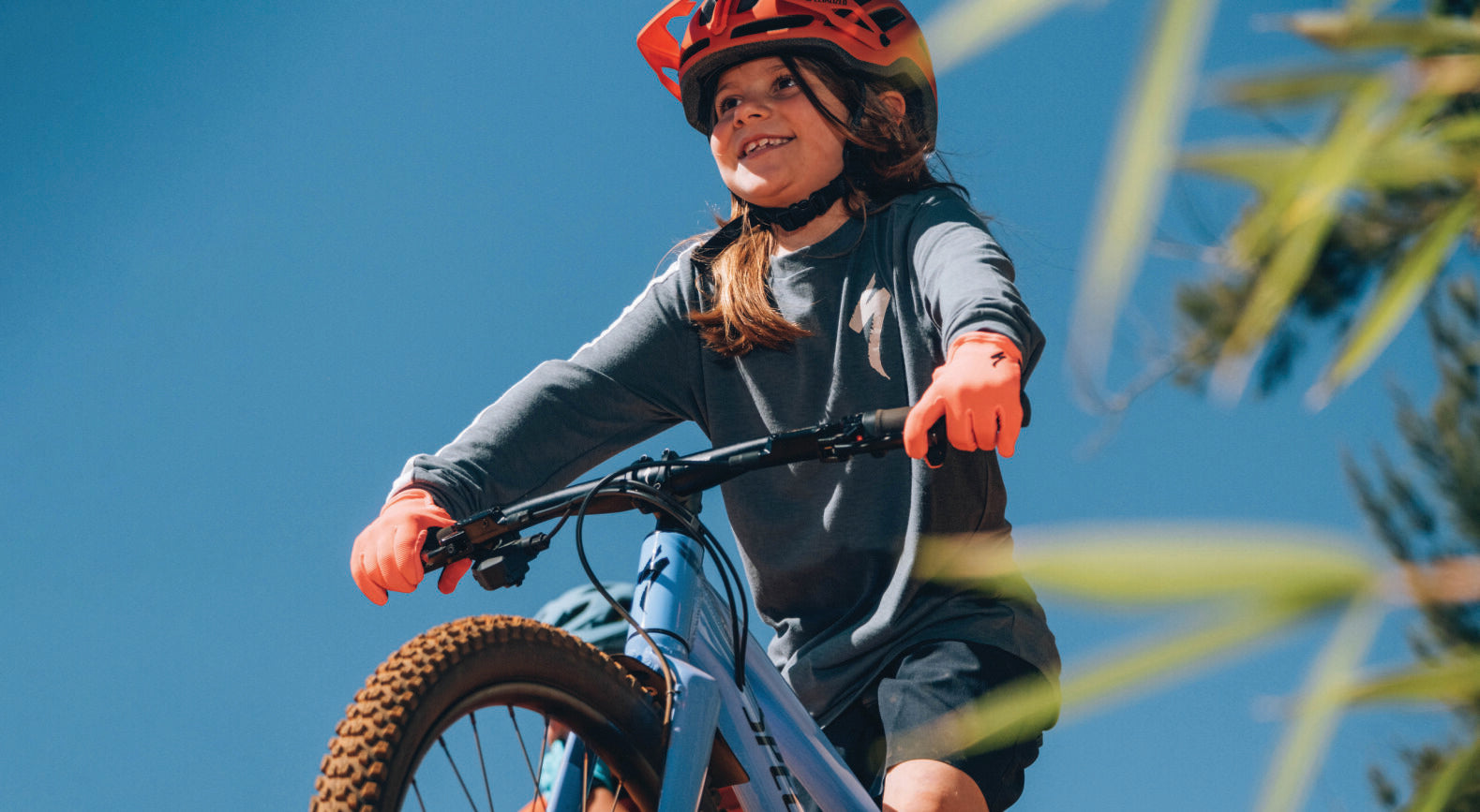 Kind fährt Specialized-Kinderfahrrad mit orangem Helm und Handschuhen, blauer Himmel im Hintergrund, für junge Radfahrer gedacht.