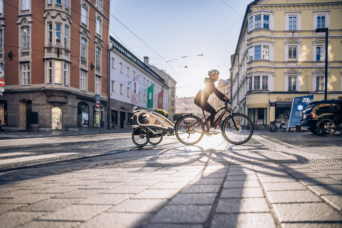 Person auf E-Bike mit Anhänger fährt bei Sonnenuntergang durch die Stadt, umgeben von historischen Gebäuden.