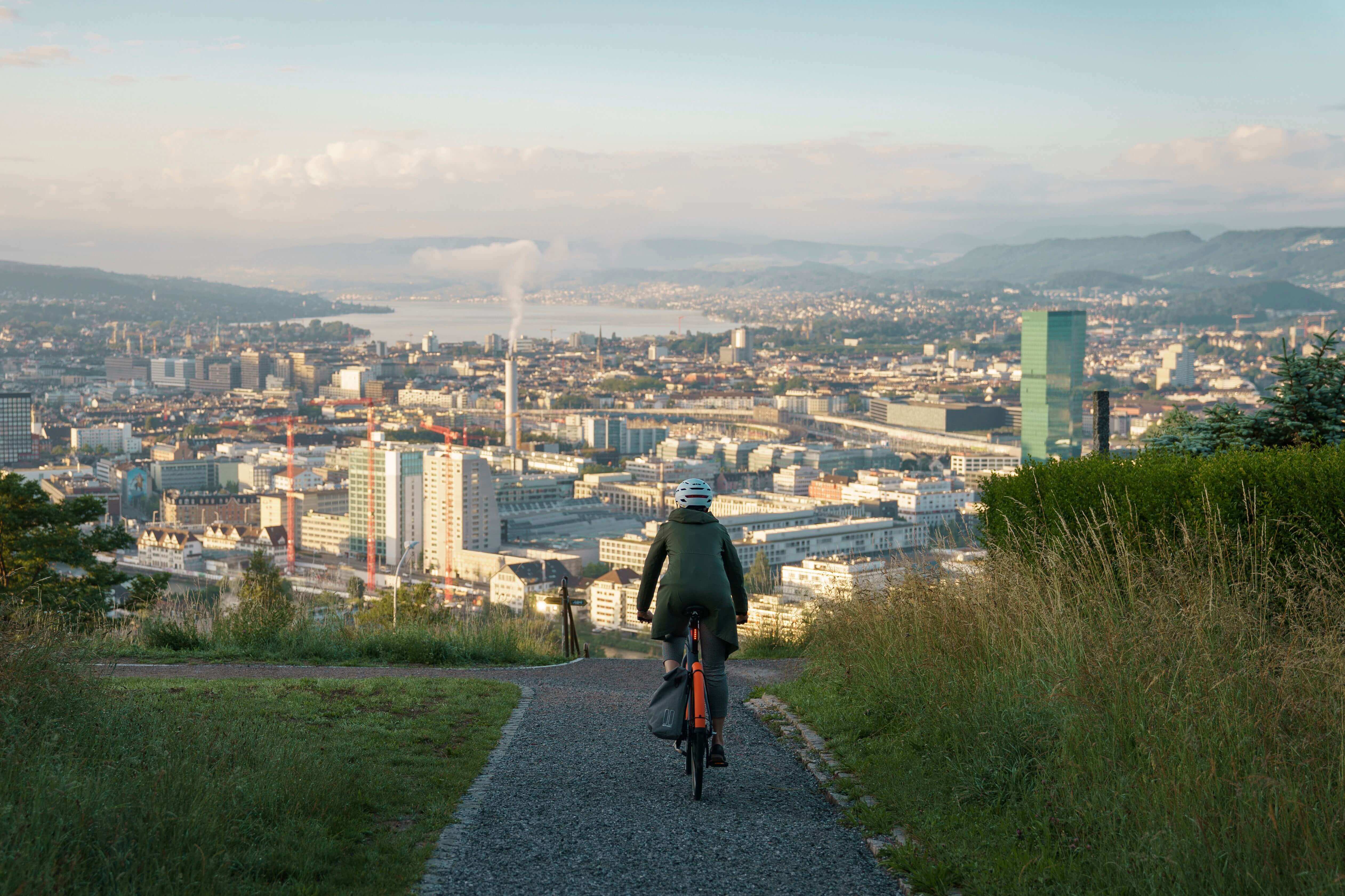 Eine Person fährt auf einem orangefarbenen Fahrrad mit Helm und Tasche eine schmale Straße hinunter, in Richtung Stadtlandschaft.