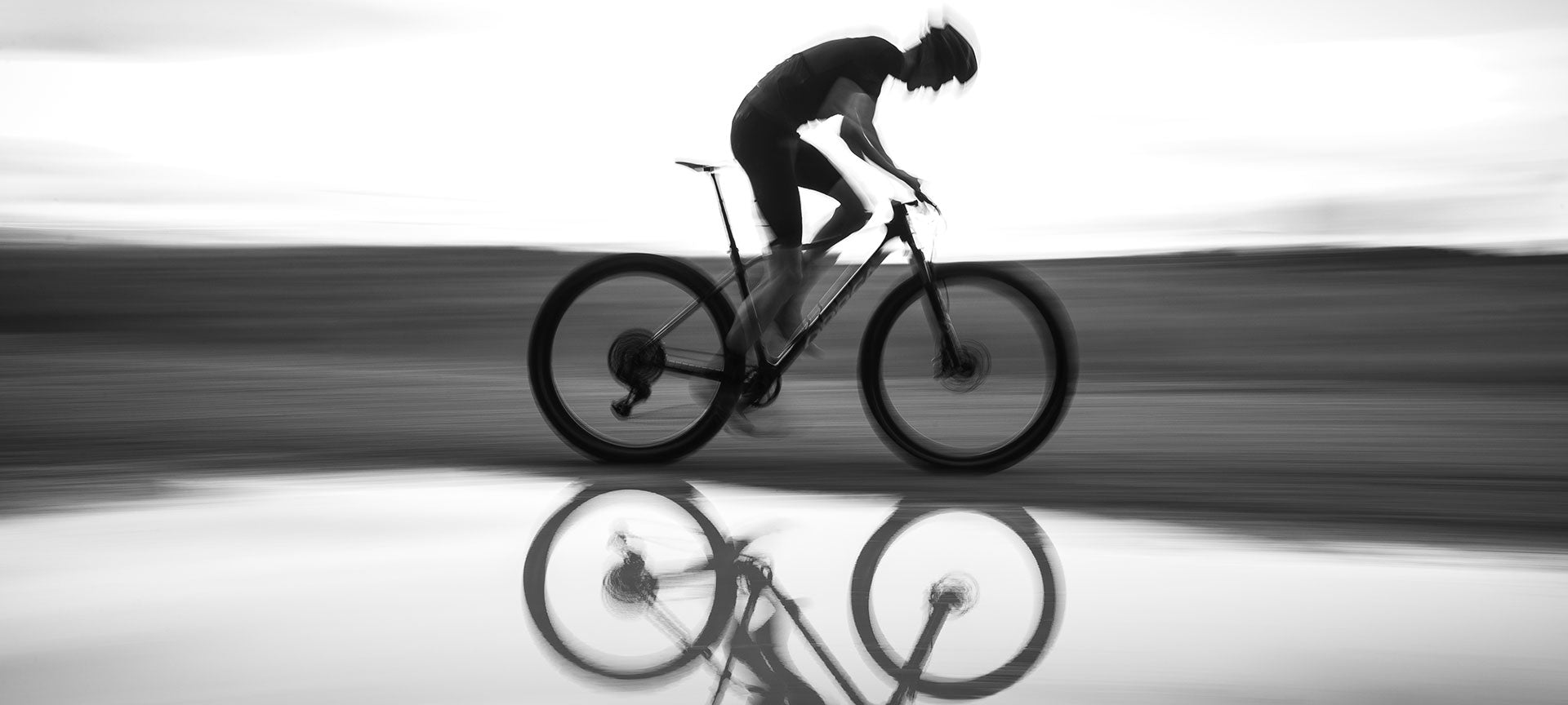 Radfahrer auf Mountainbike Orbea Alma, dynamische Fahrt über Wasserpfütze, im Hintergrund verschwommene Landschaft.