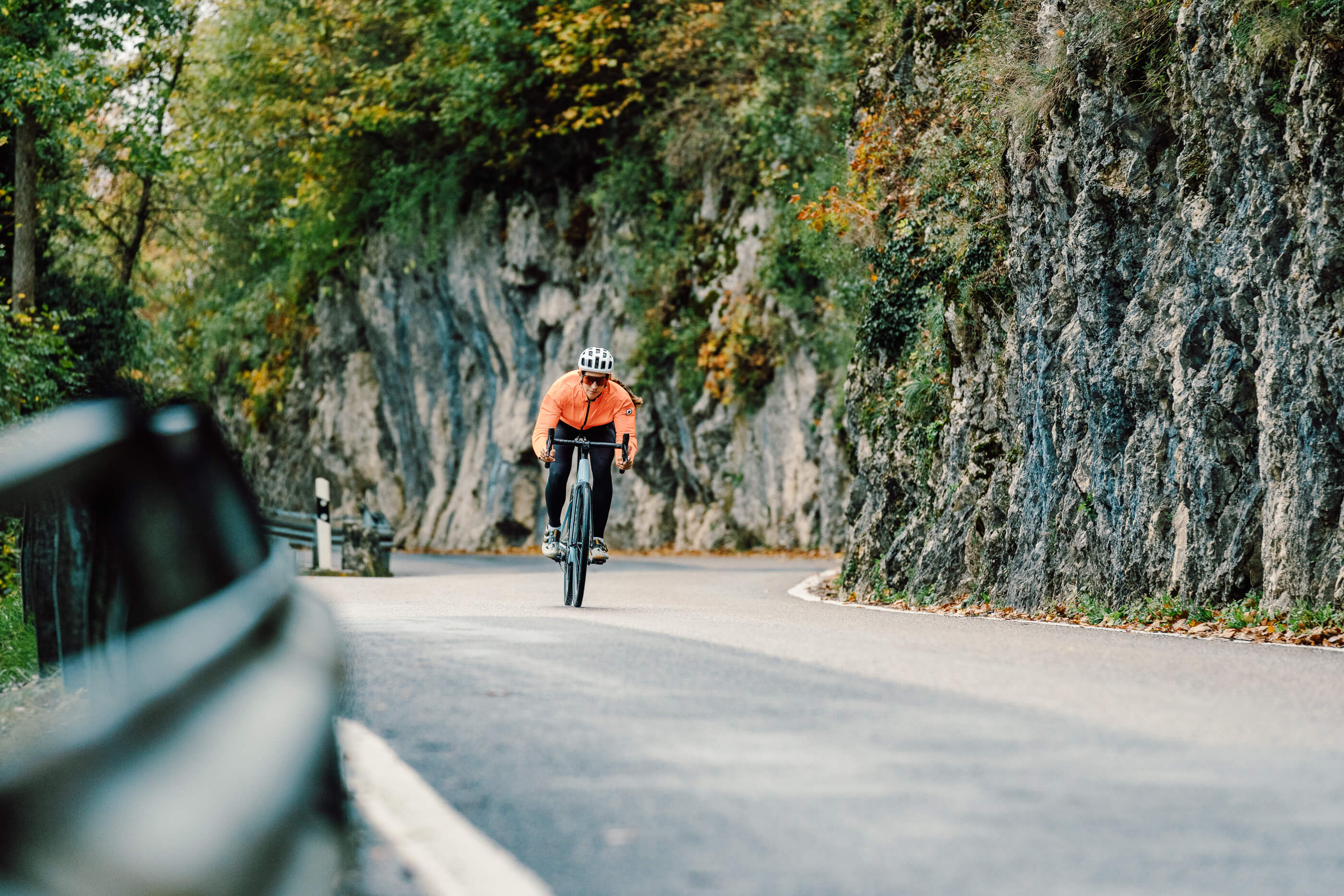Radfahrer in oranger Jacke fährt auf einem Rennrad durch eine kurvenreiche Straße in bergiger Landschaft.