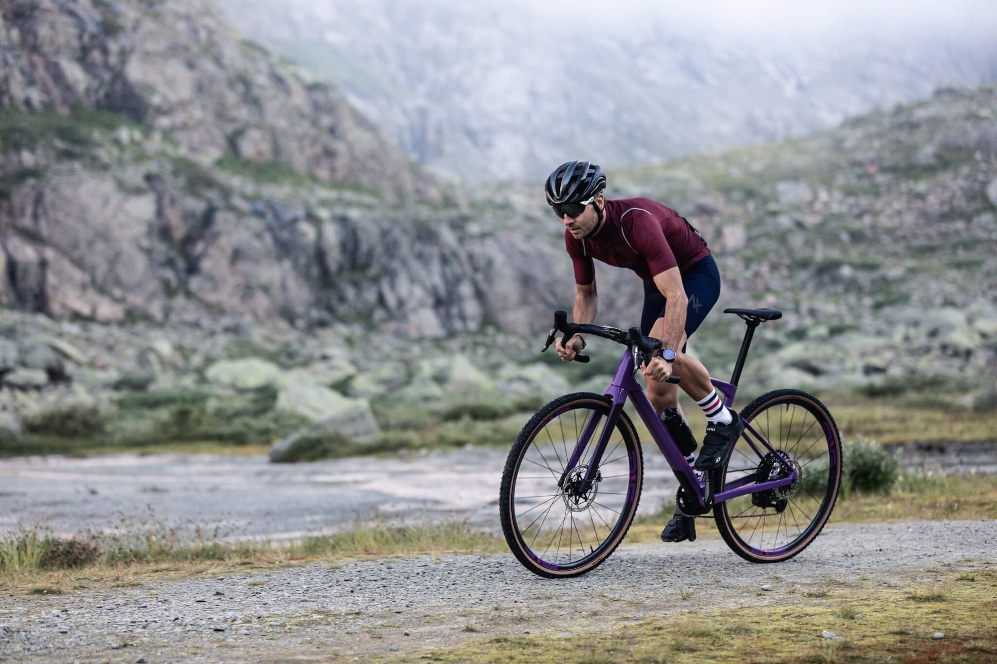Mann fährt ein lila BMC URS Gravelbike auf einem steinigen Weg in einer bergigen Landschaft.
