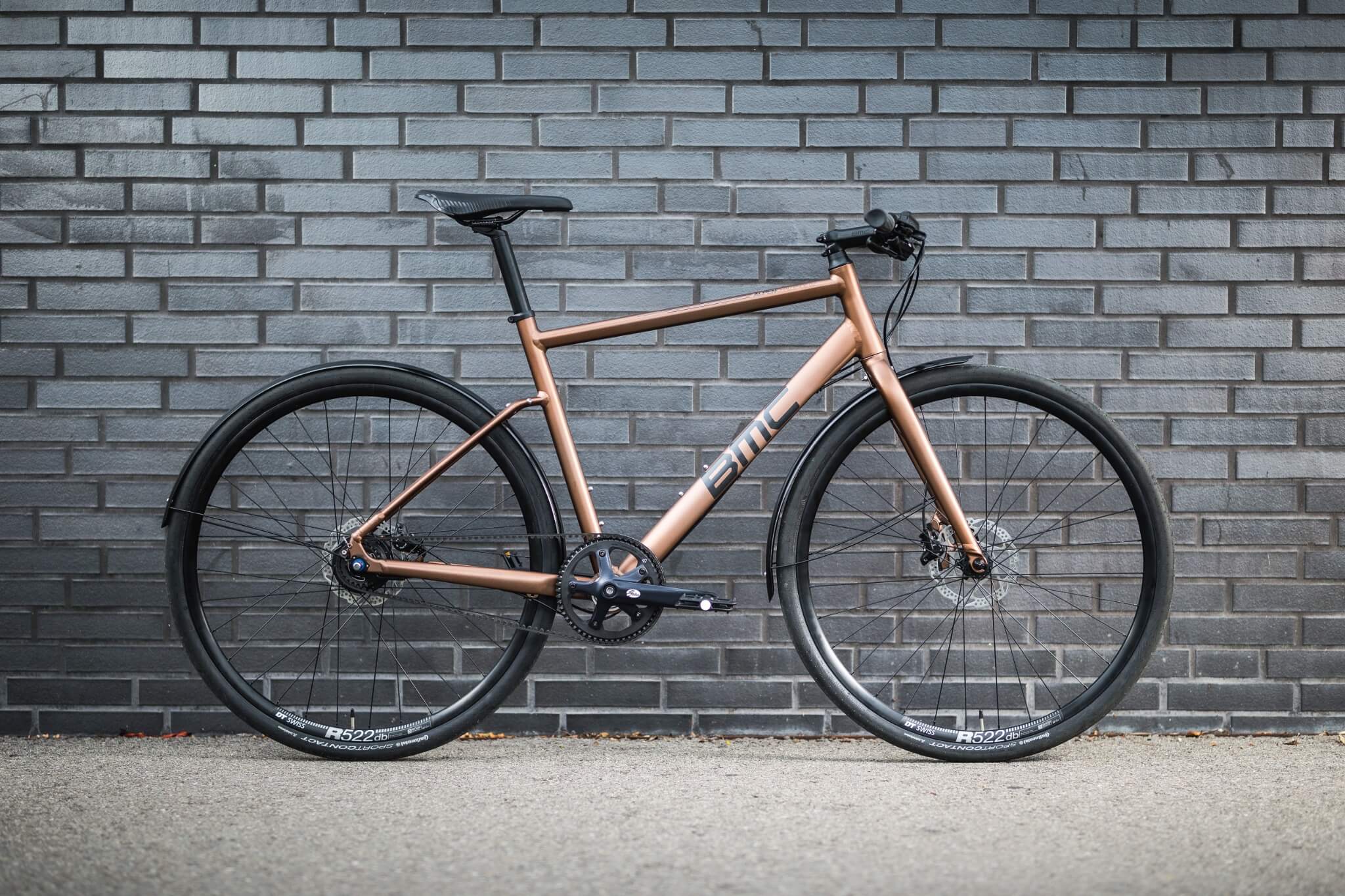 Kupferfarbenes BMC Citybike vor grauer Ziegelwand; sportliches Design, ideal für urbane Pendler.