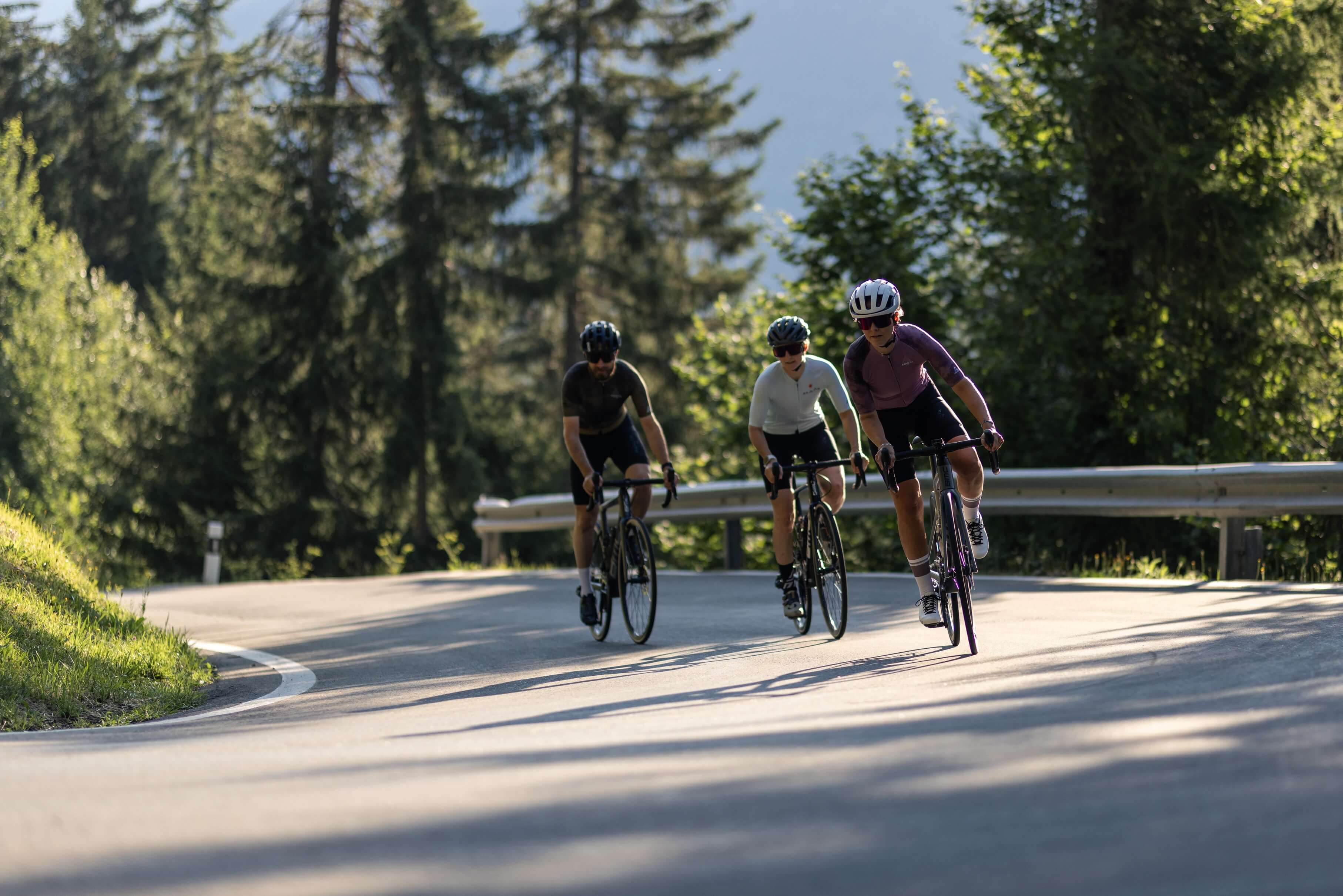 Drei Radfahrer auf einer Bergstraße, umgeben von Wald, bei Sonnenlicht, keine speziellen Marken erkennbar.