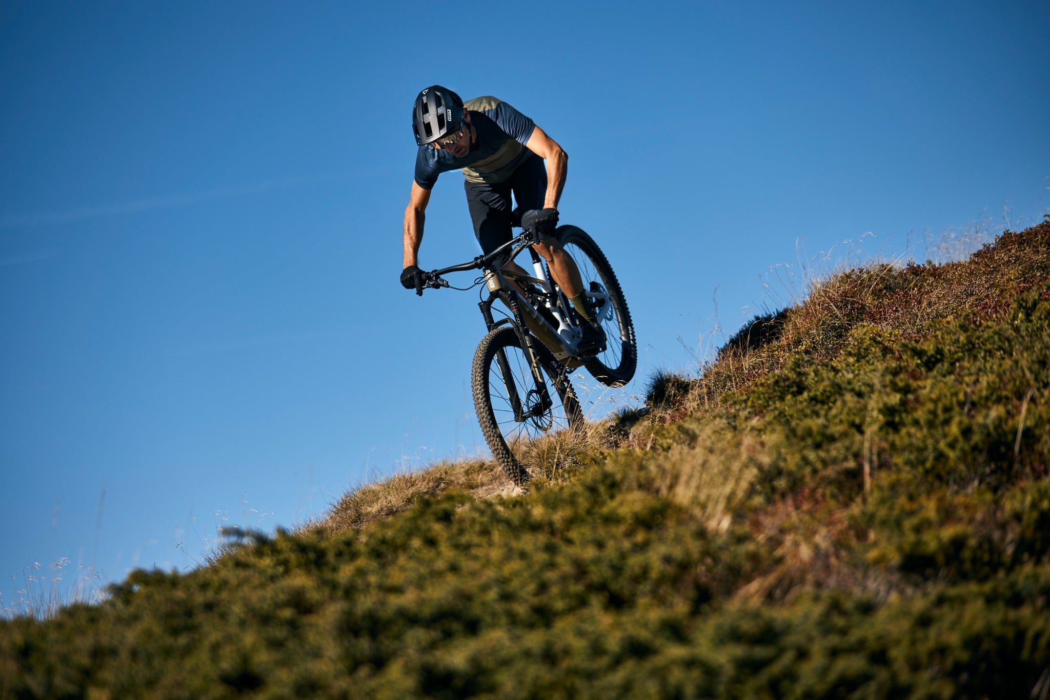 Mountainbiker in Aktion auf einem BMC Fourstroke AMP, steile Abfahrt auf einer Grasböschung bei blauem Himmel.