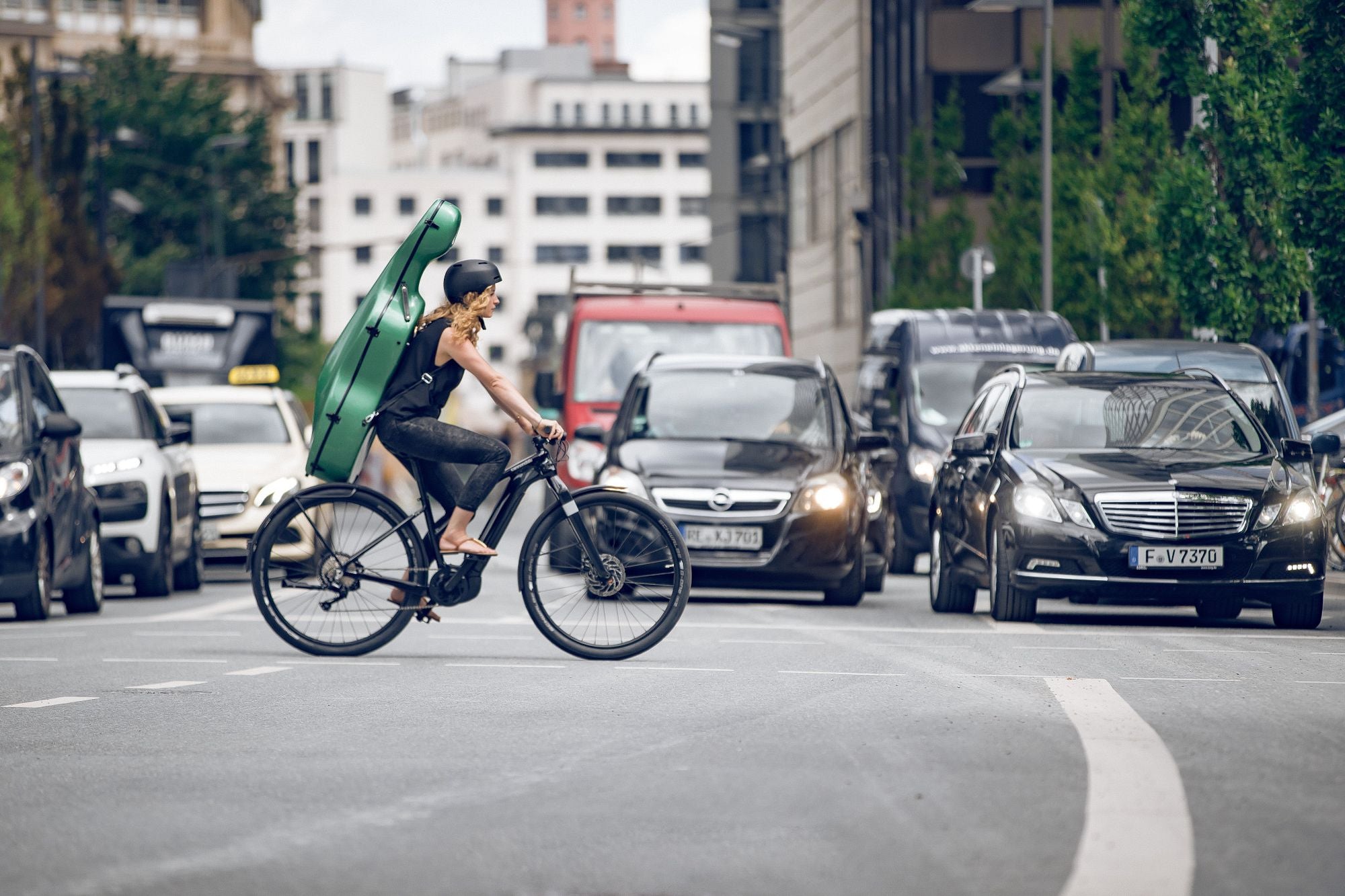 Radfahrerin mit grünem Instrumentenkoffer fährt Cannondale E-Bike durch belebte Stadtstraße.