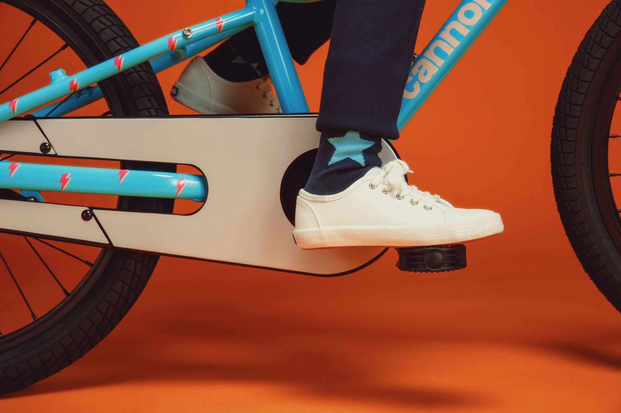 Kind auf Cannondale-Kinderfahrrad, blau mit Blitzmuster, weißer Schuh auf Pedal, oranger Hintergrund.