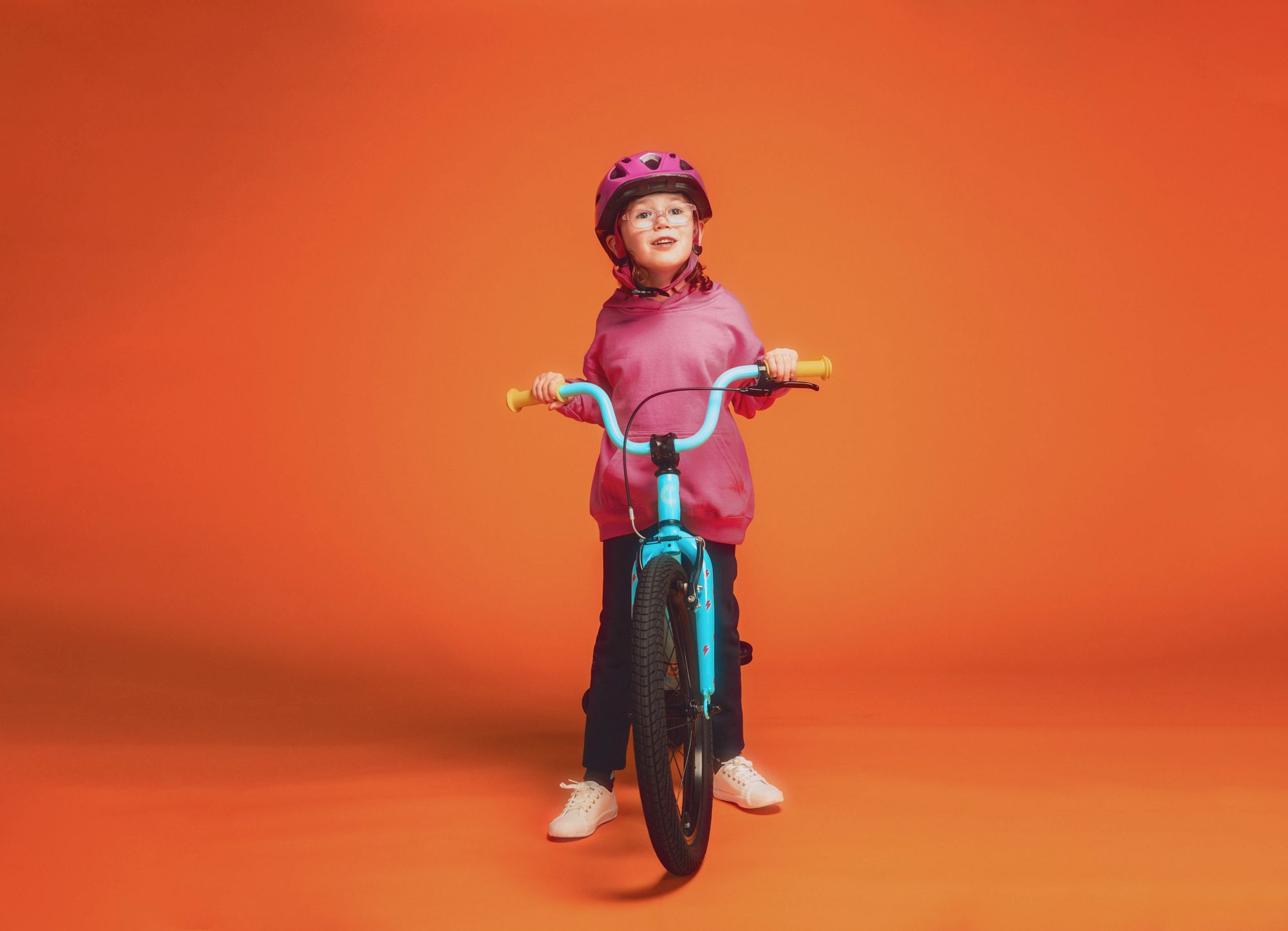 Kind mit pinkem Helm und Pulli auf einem hellblauen Cannondale-Kinderfahrrad vor orangefarbenem Hintergrund.