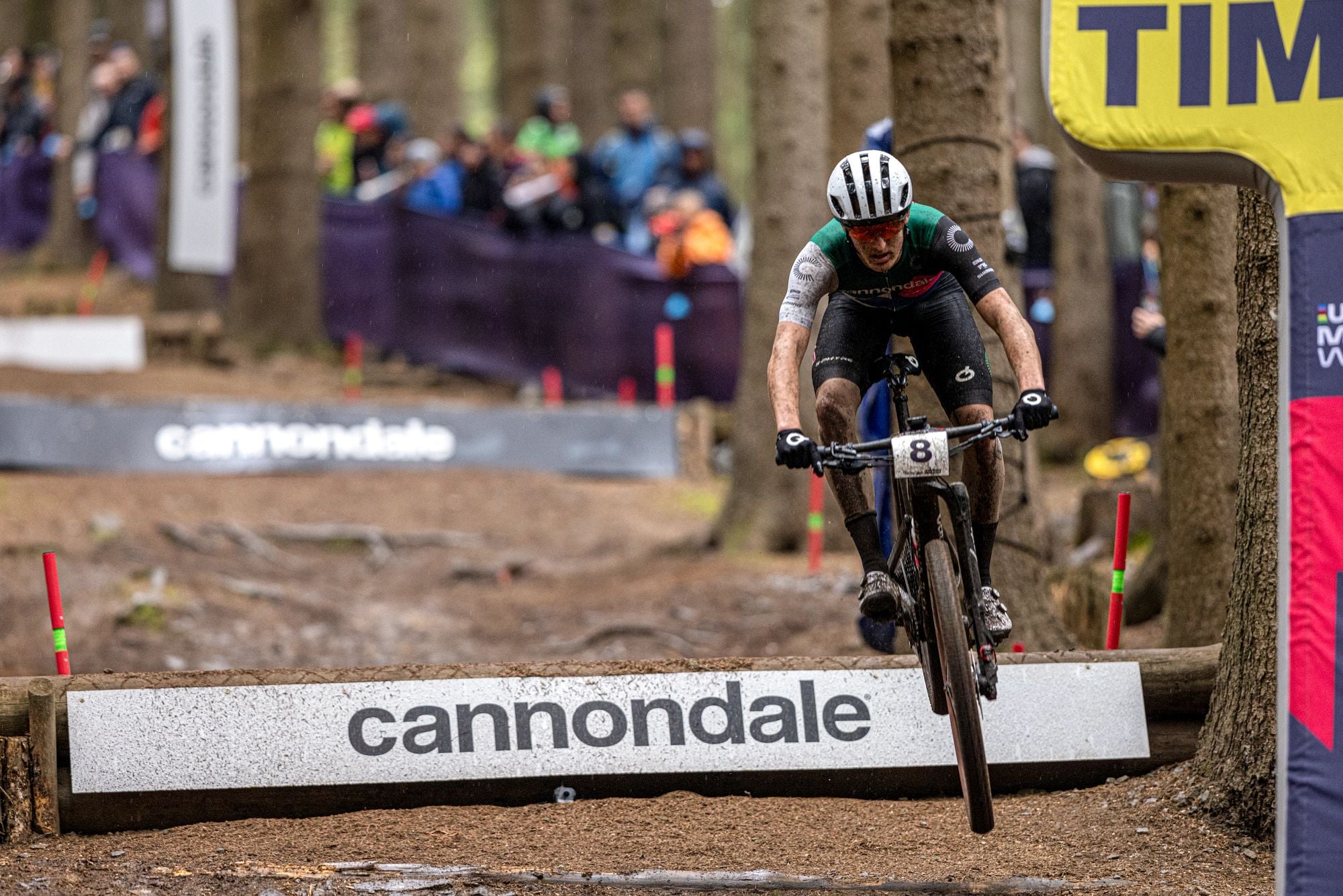 Mountainbiker springt bei Rennen im Wald über Hindernis mit Cannondale-Schriftzug, umgeben von Zuschauern.