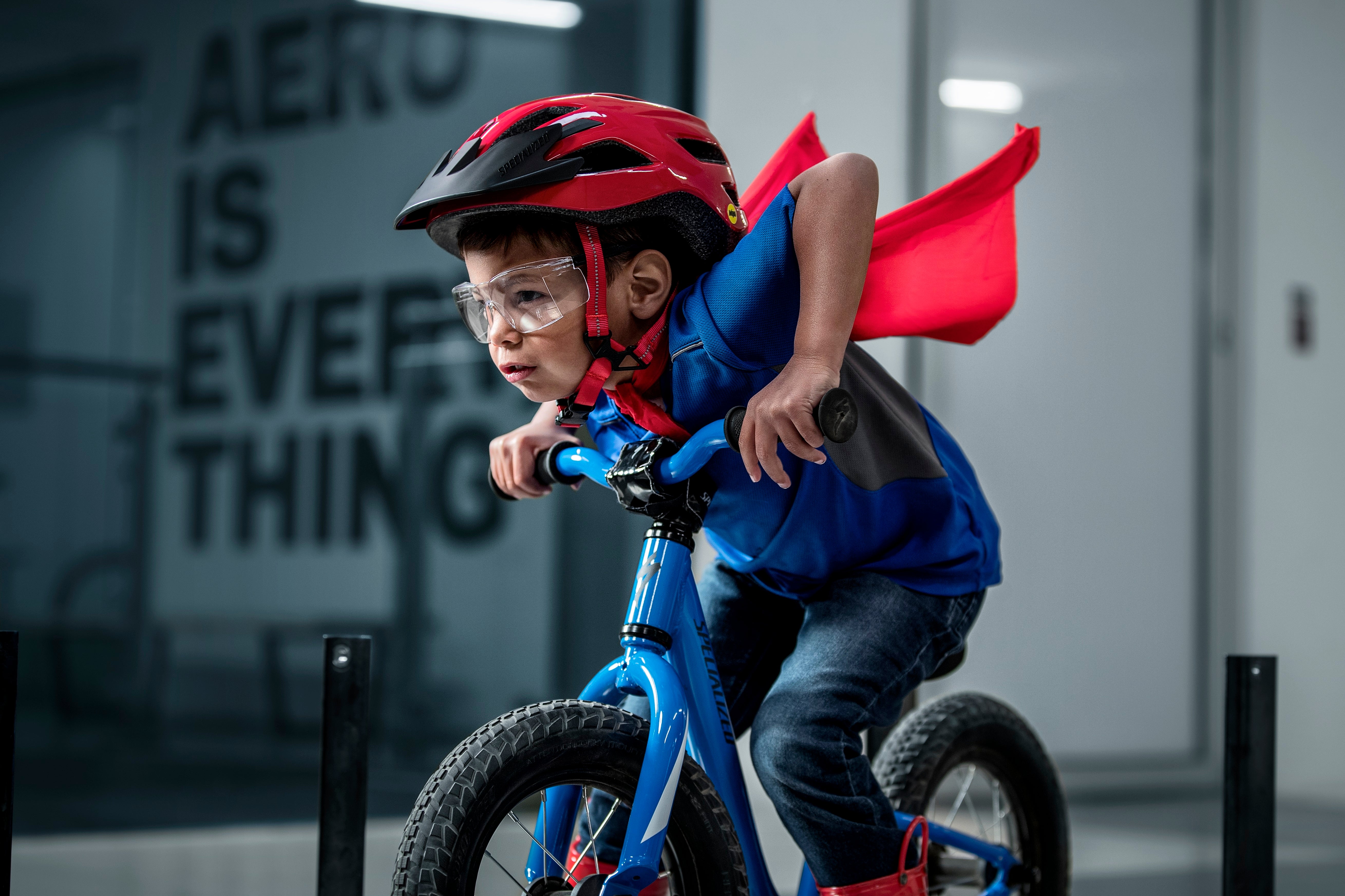 Kind auf Specialized Laufrad mit rotem Helm und Umhang, trägt Schutzbrille, indoor Rennszene.