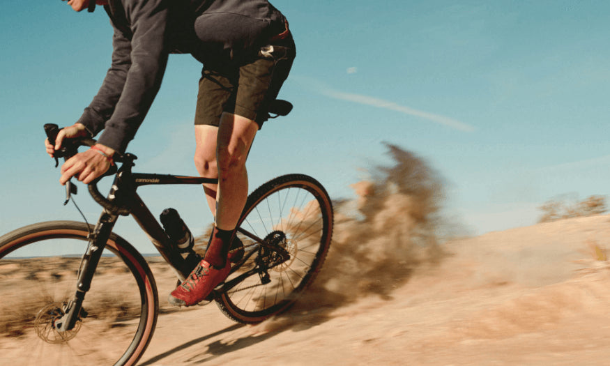 Person fährt schnell auf einem Cannondale Gravelbike über eine staubige Piste unter blauem Himmel.