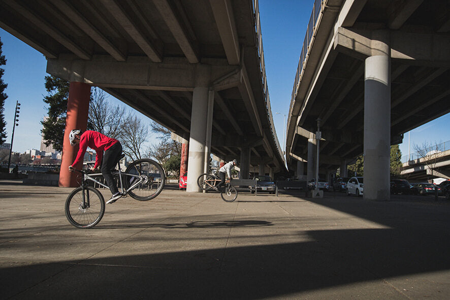 Zwei Personen auf Specialized Hardtail-Mountainbikes machen Stunts unter einer Stadtbrücke.