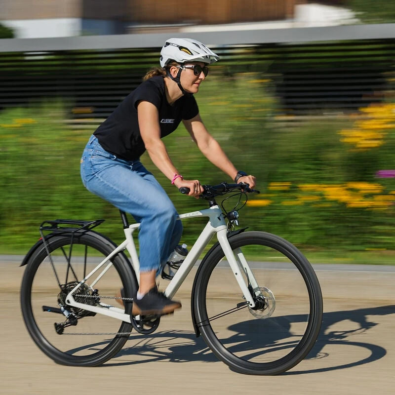 Person auf einem weißen Cube Gravelbike trägt Helm und Sonnenbrille, fährt draußen auf einer Straße.