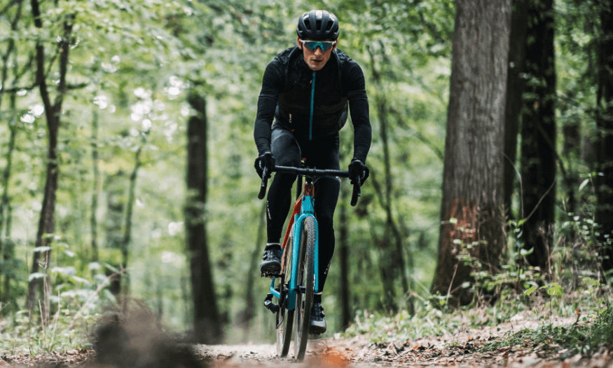 Radfahrer auf einem blauen Gravelbike im Wald, trägt Helm und schwarze Sportkleidung.