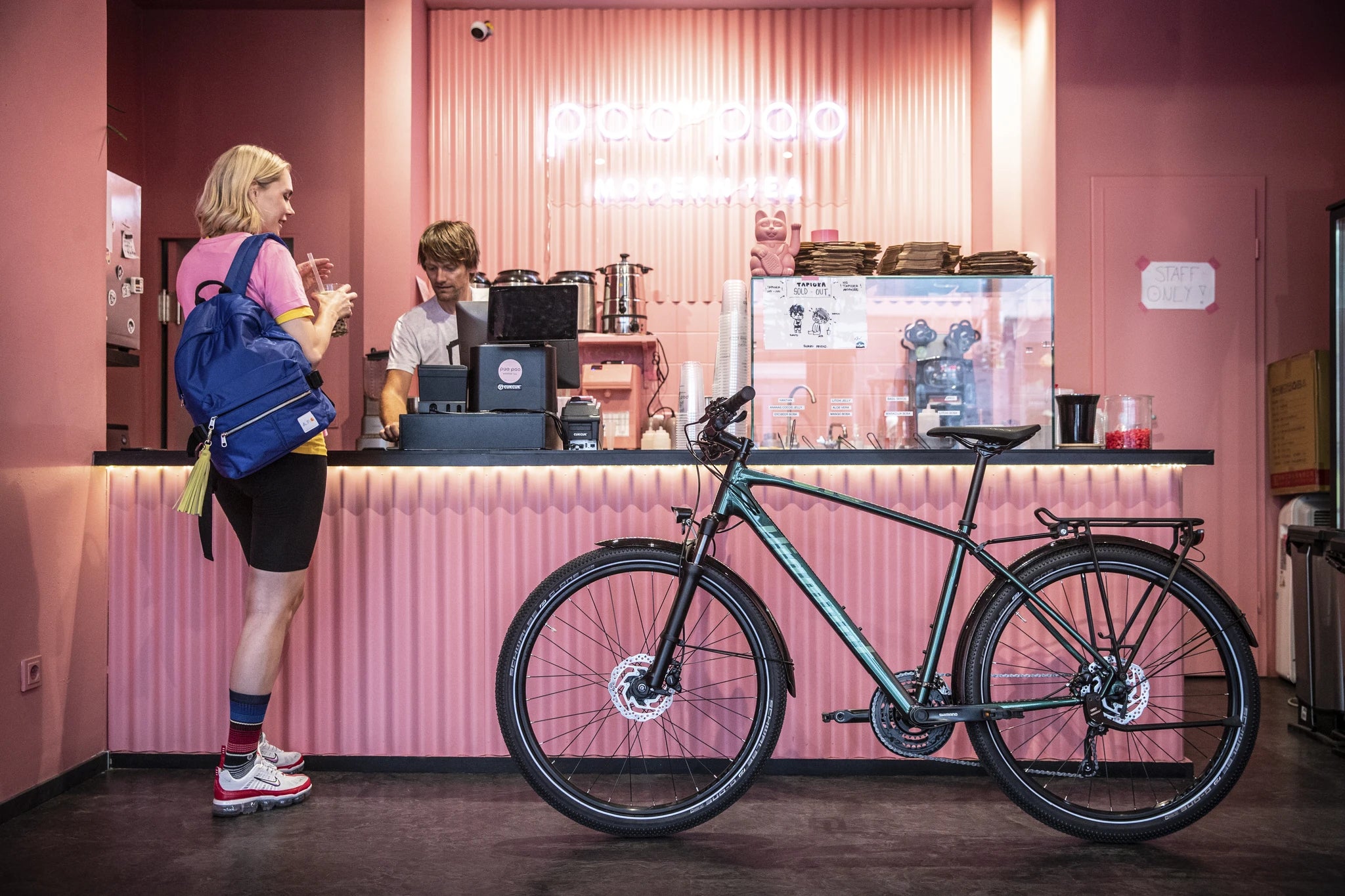 Frau mit blauem Rucksack im Bubble Tea Café, grünes Fahrrad vorne, Barista bedient hinter pinker Theke.