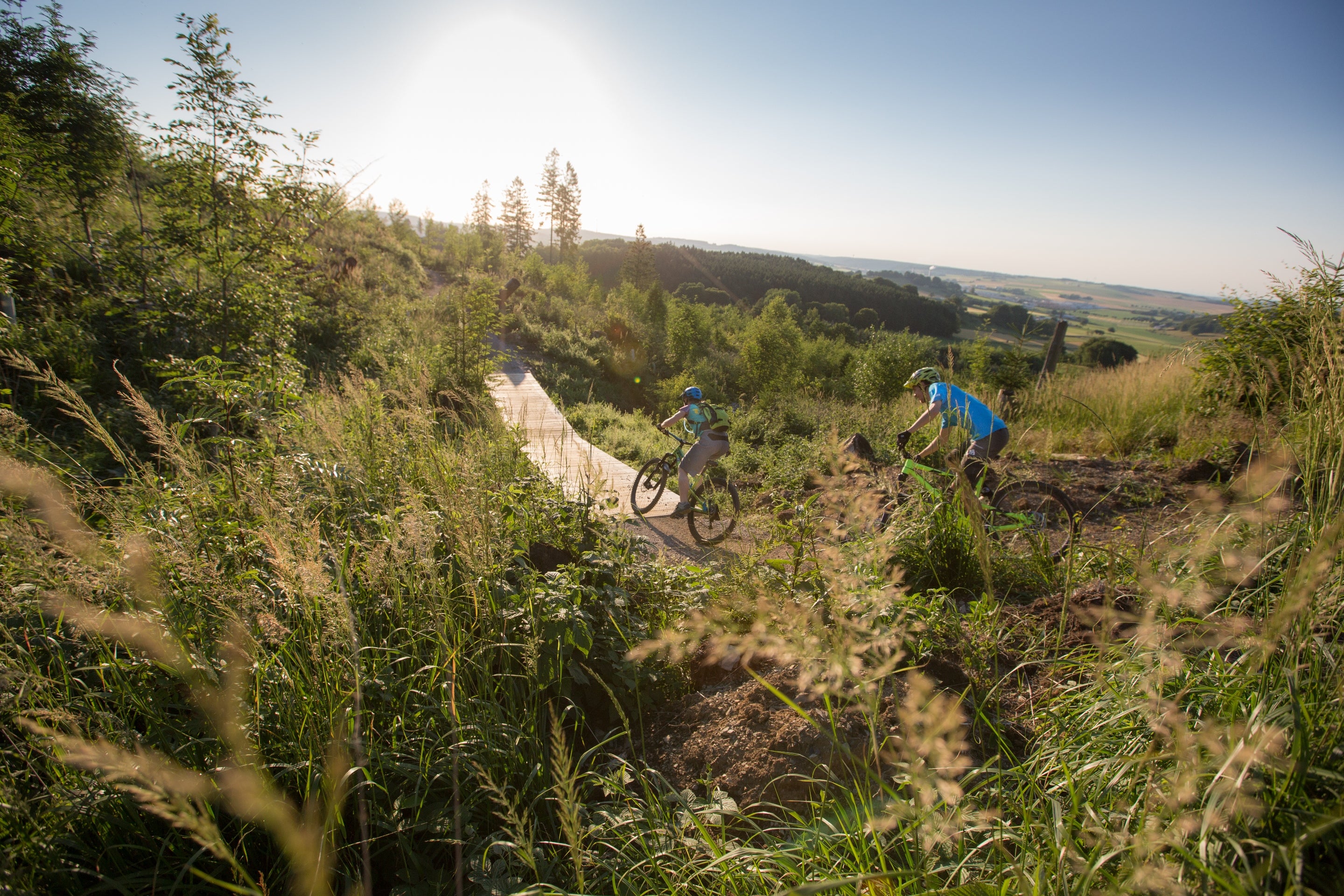 Zwei Mountainbiker fahren auf einem Pfad in hügeliger, bewaldeter Landschaft bei Sonnenuntergang.