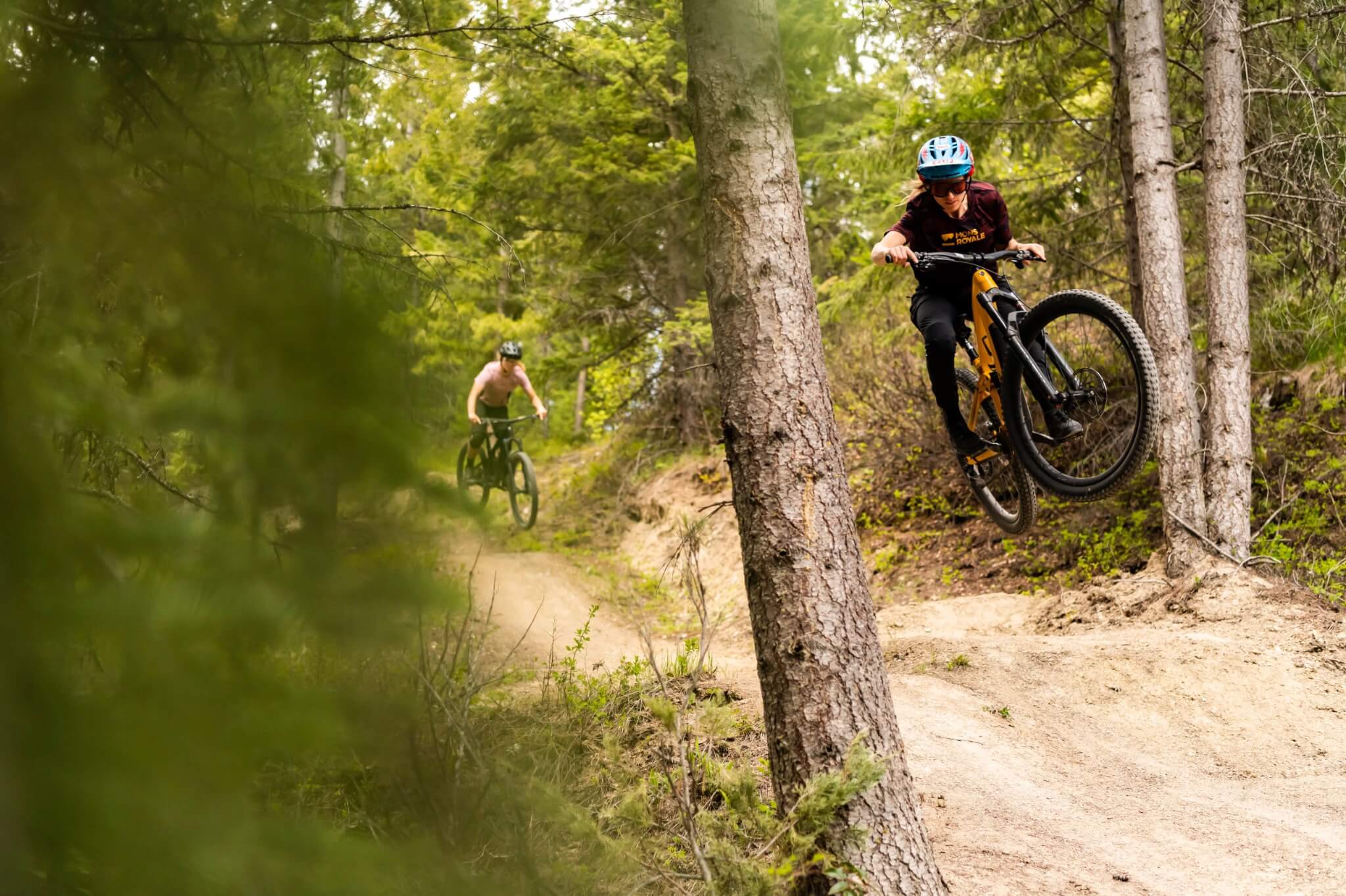 Zwei Mountainbiker fahren durch einen Waldpfad, einer springt mit einem Trek Fuel EX Fahrrad in die Luft.