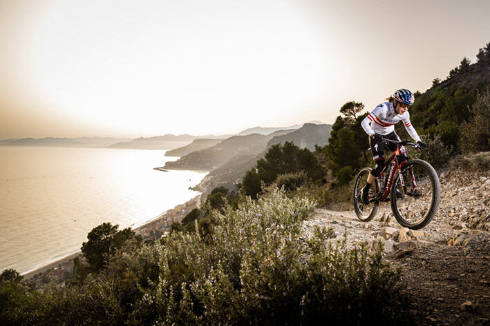 Mountainbiker auf einem Specialized Fully fährt auf einem Küstenpfad bei Sonnenuntergang. Meer und Berge im Hintergrund.