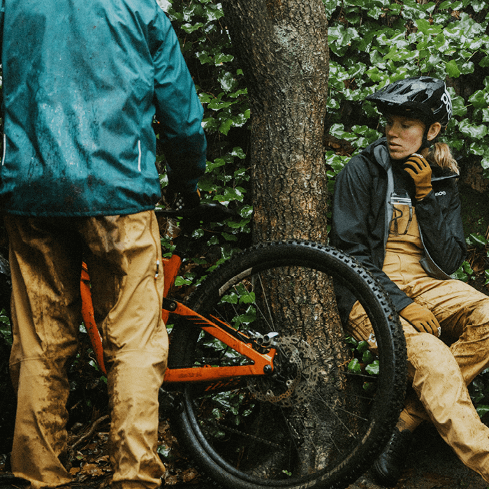 Zwei Personen in Outdoor-Bekleidung mit Mountainbike im Wald, eine Person sitzt, die andere steht am Bike.