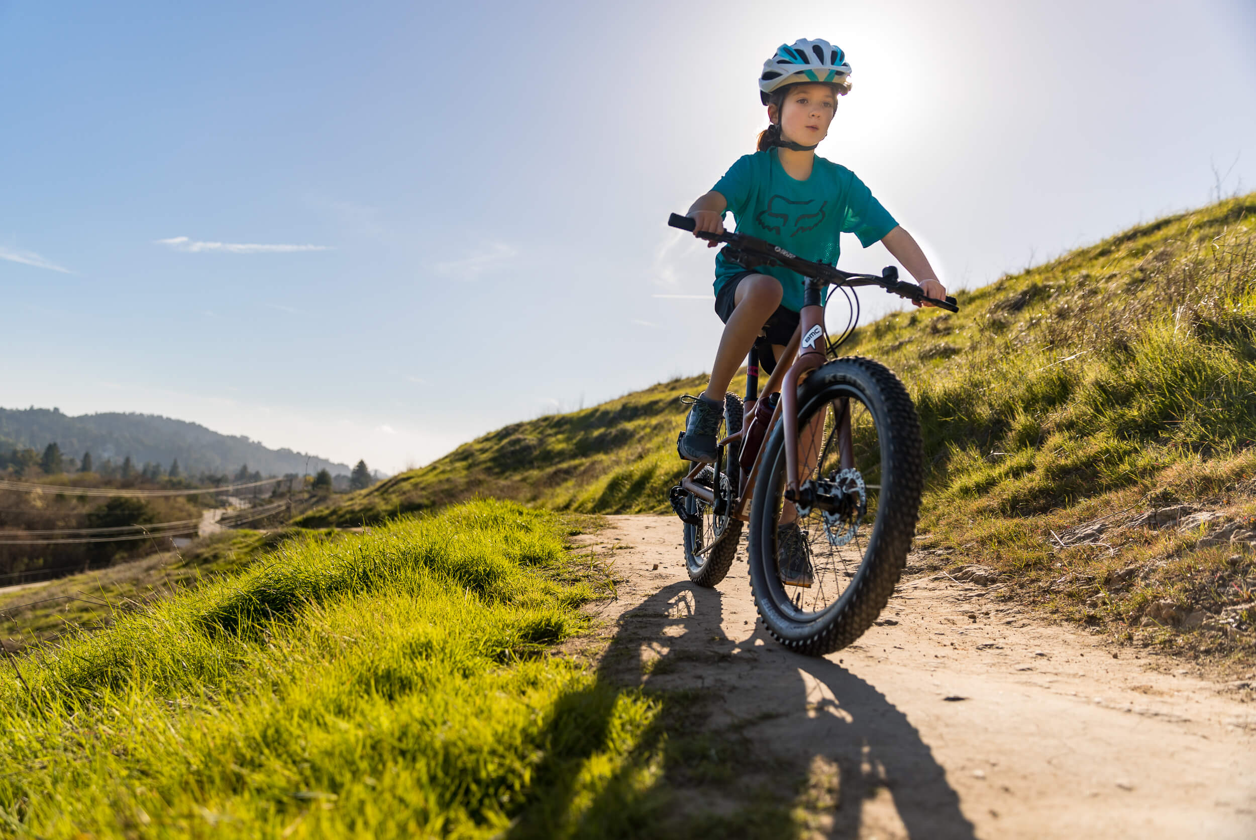 Kind mit Helm auf Mountainbike fährt auf sonnigem Pfad durch hügelige Landschaft; geeignet für junge Abenteurer.