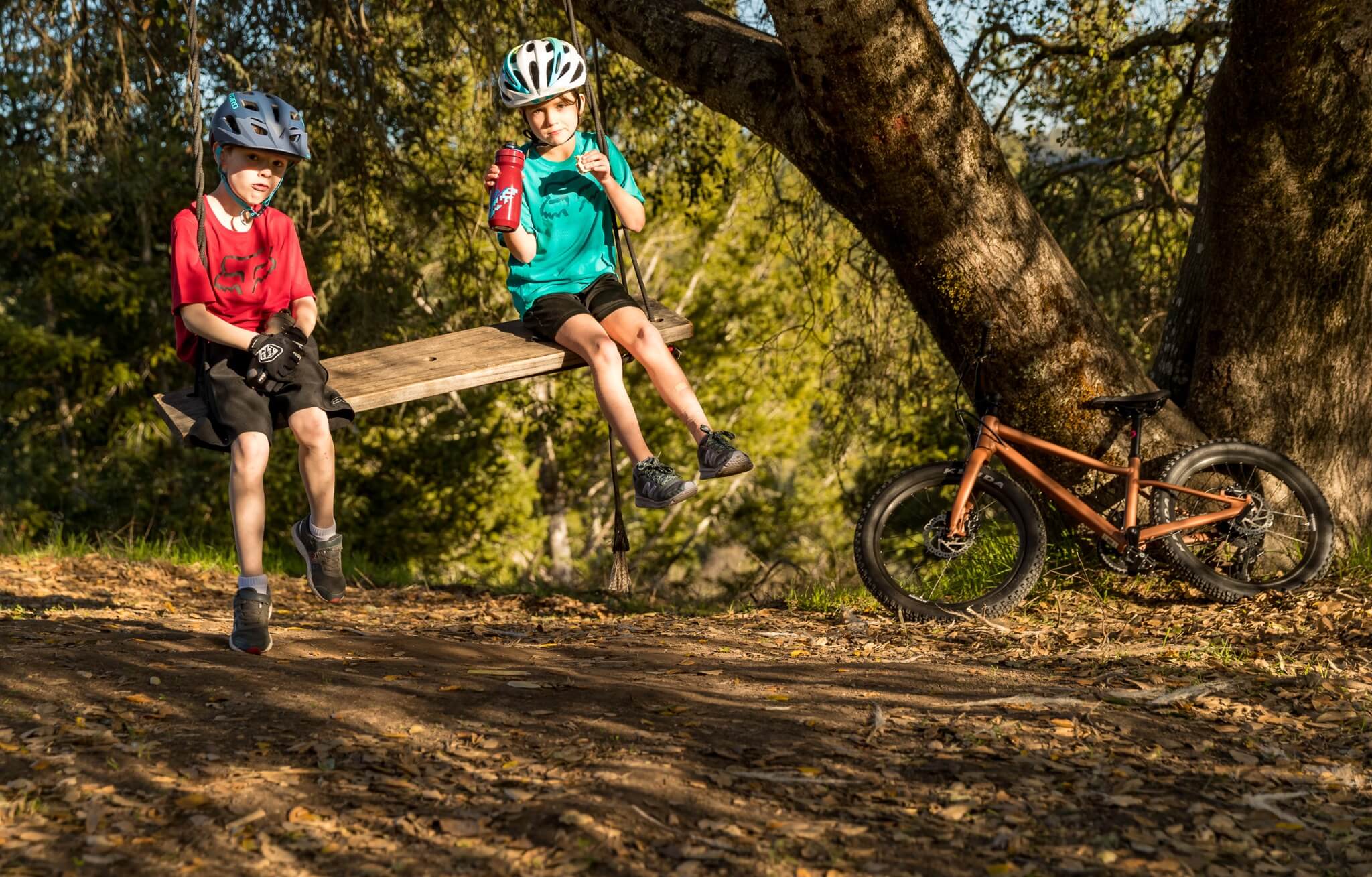 Zwei Kinder auf einer Schaukel im Wald, tragen Helme und bunte Trikots, ein Fahrrad lehnt am Baum.
