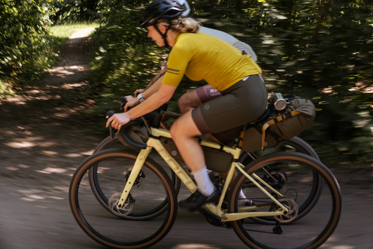 Zwei Radfahrer auf gelbem Gravelbike mit Taschen, FOCUS-Collection, fahren durch einen Wald.
