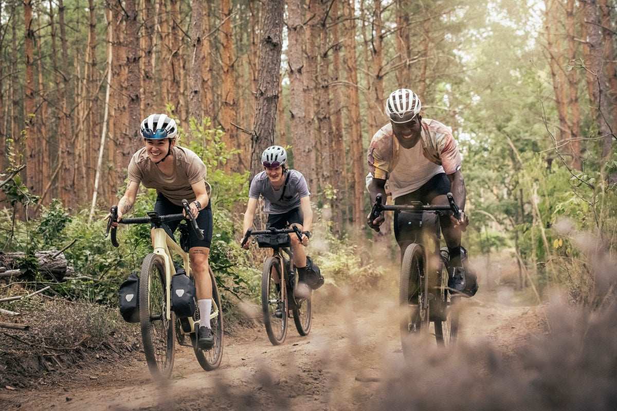 Drei Radfahrer in einem Wald auf Gravelbikes, lächelnd und fortschreitend, tragen Helme und Casual-Bekleidung.