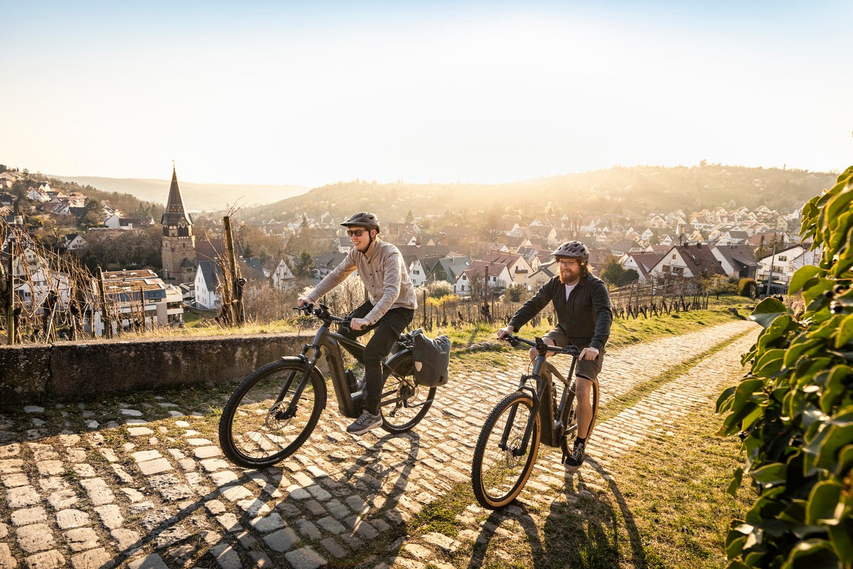 Zwei Personen auf Fahrrädern in einer ländlichen, hügeligen Gegend bei Sonnenuntergang.