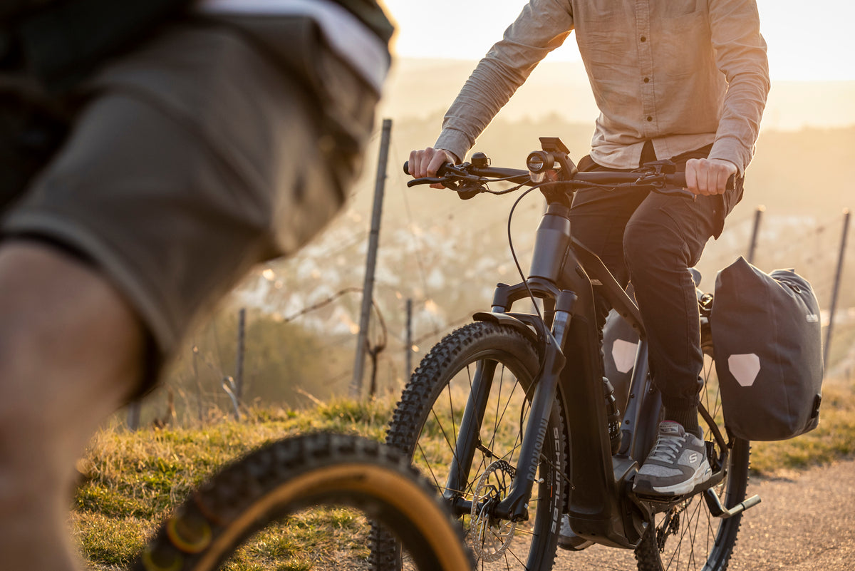 Zwei Personen fahren bei Sonnenuntergang auf einem Focus E-Bike mit Satteltaschen auf einem Feldweg.