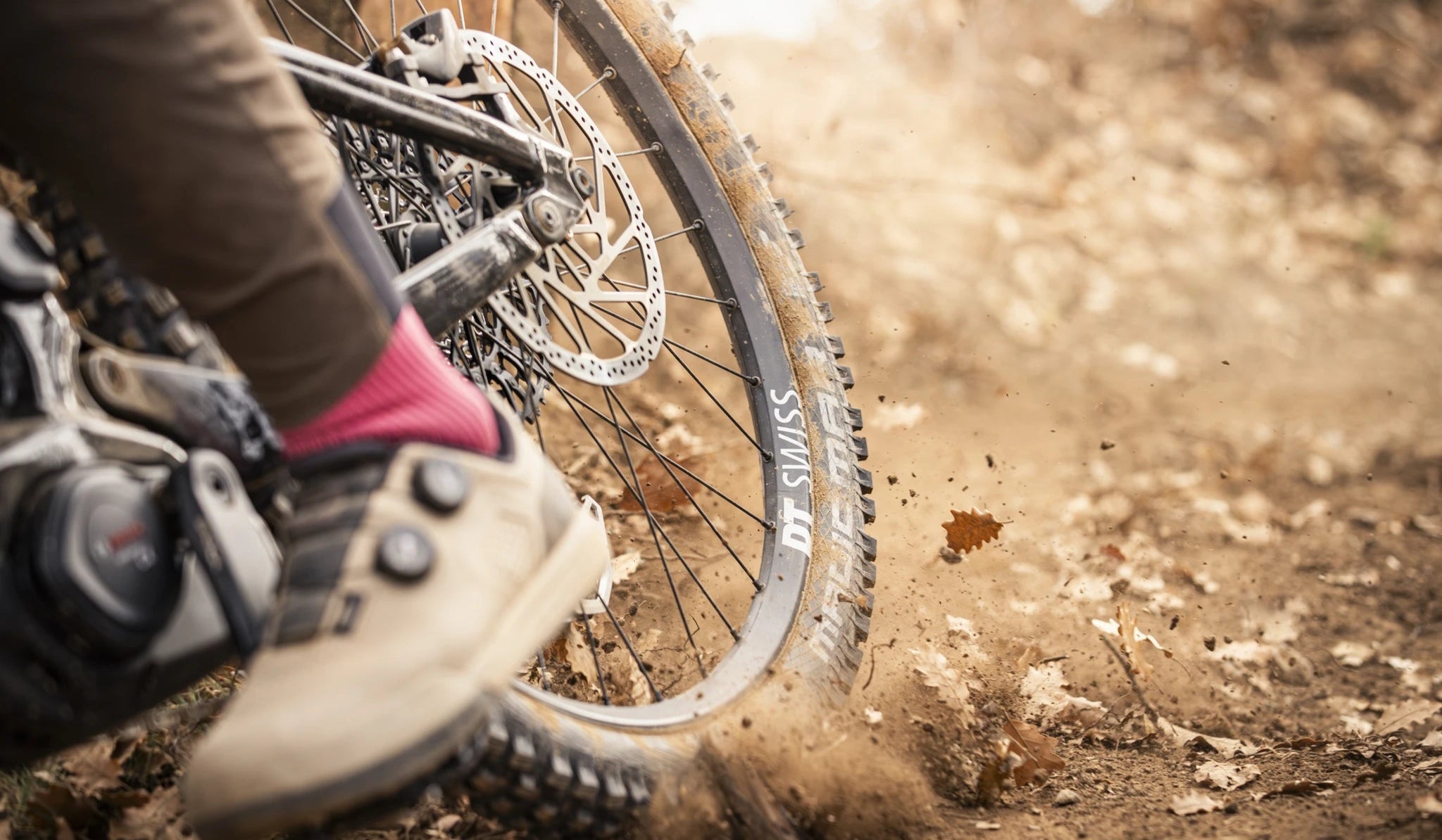 Nahaufnahme eines Mountainbikes in Aktion, DT Swiss Reifen, Fahrer mit pinken Socken und hellen Schuhen im Gelände.