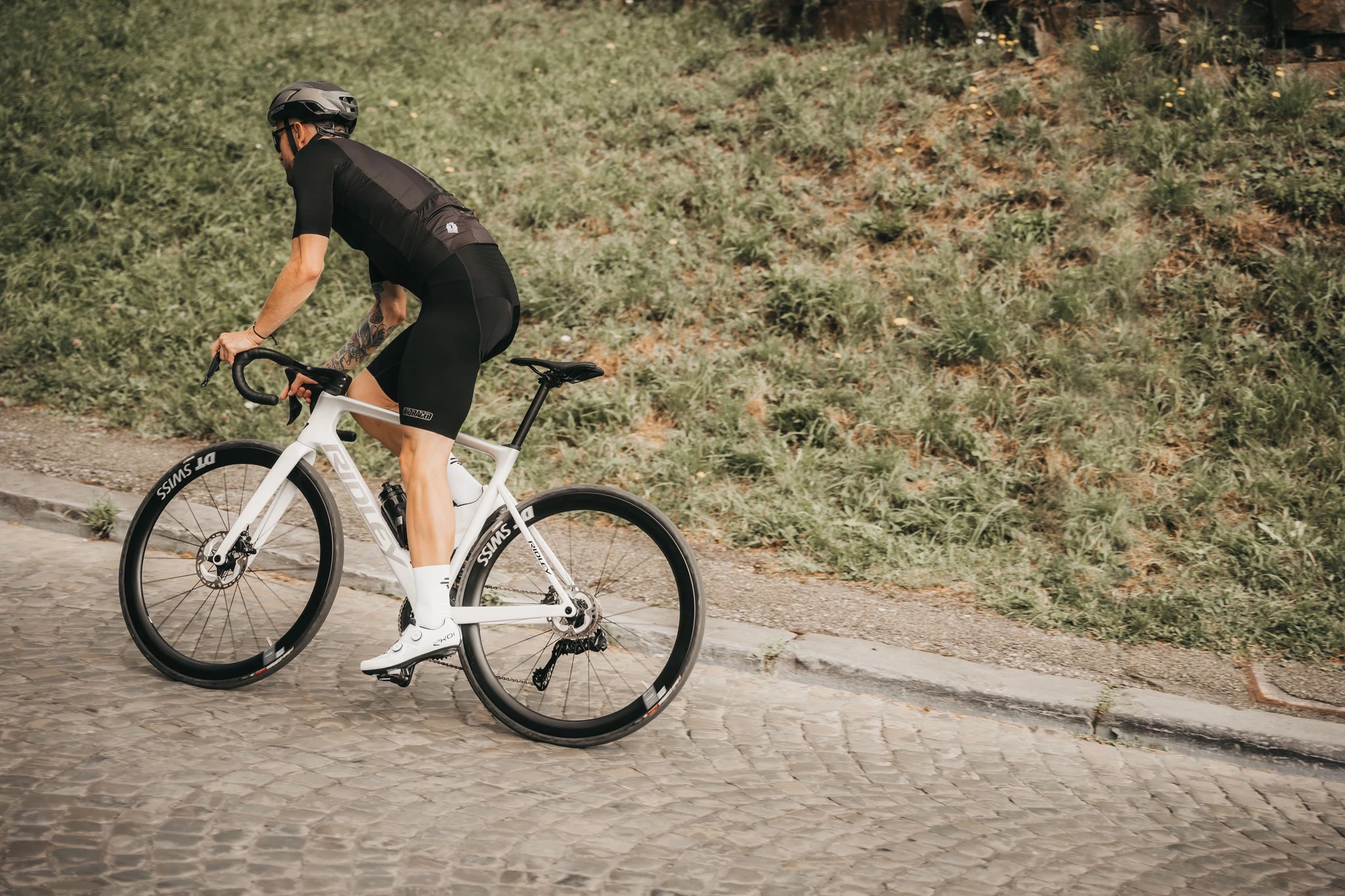 Radfahrer auf einem weißen Ridley Rennrad auf Kopfsteinpflaster, trägt Fahrradbekleidung und Helm, Natur im Hintergrund.