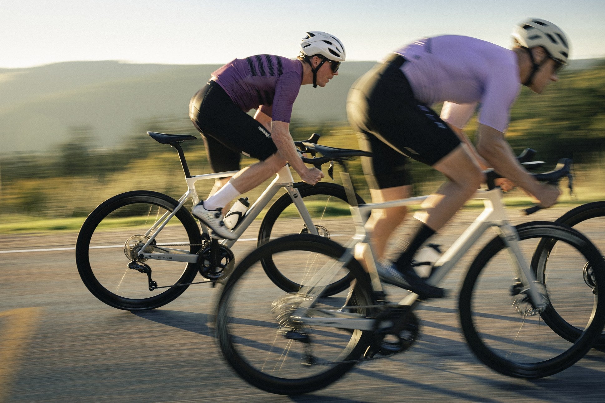 Zwei Radfahrer in Sportkleidung fahren auf Focus Izalco Max Rennrädern bei Sonnenuntergang auf einer Straße.
