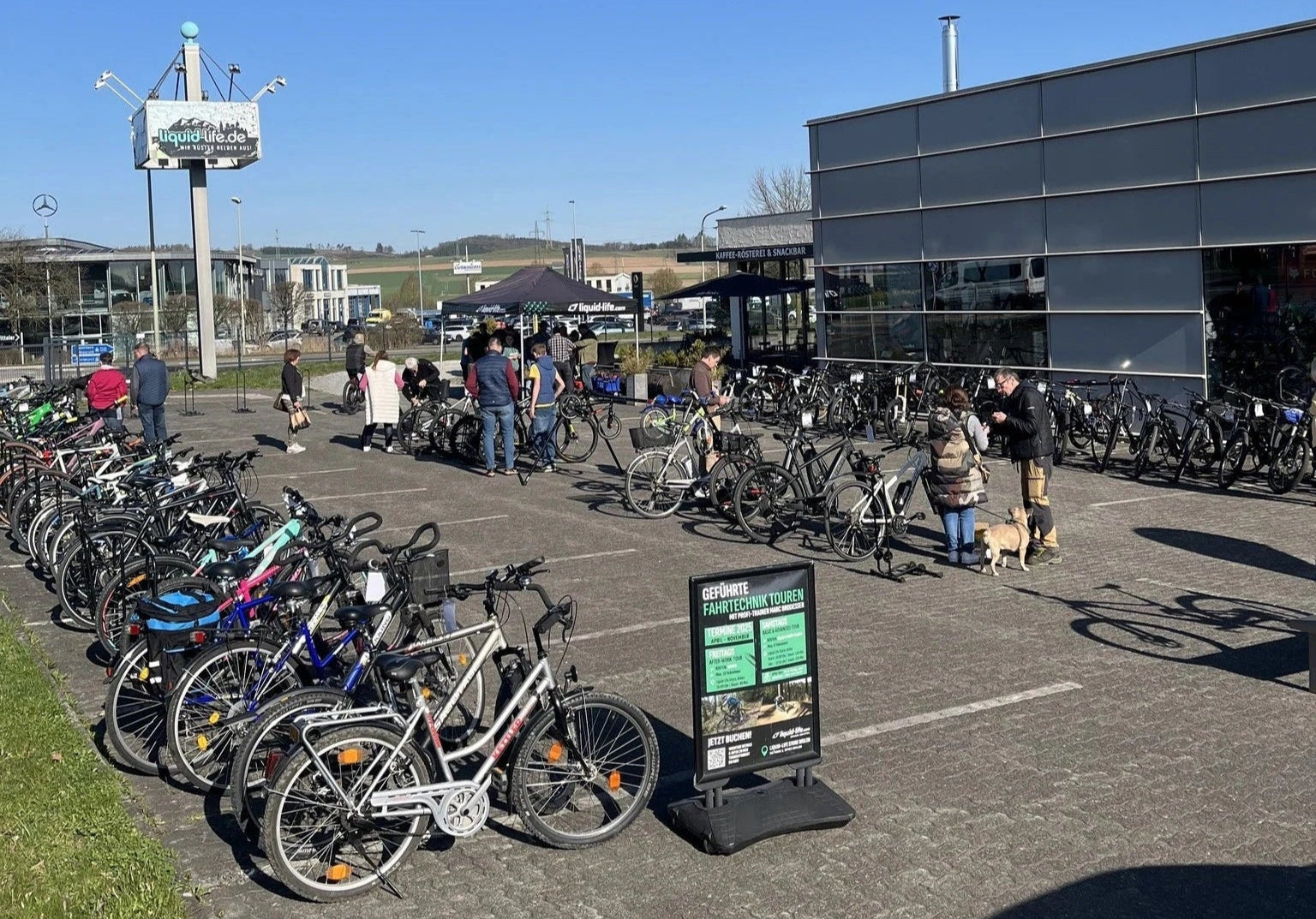 Fahrradmarkt mit zahlreichen Rädern, Menschen beim Stöbern, Schild zu Fahrtechnik-Touren, draußen bei sonnigem Wetter.