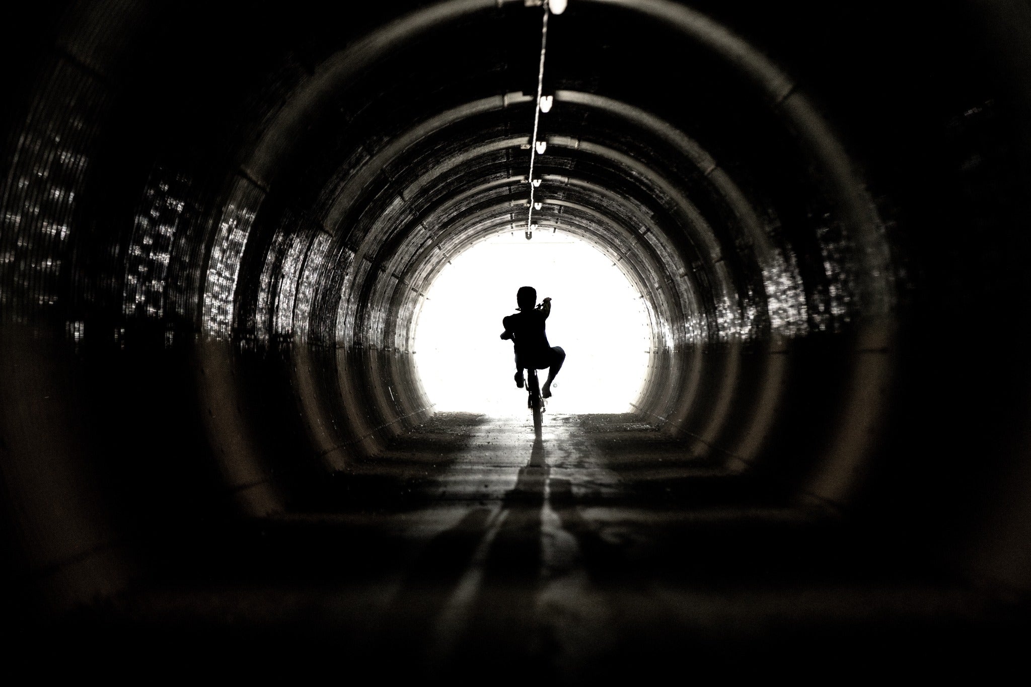 Schattenriss einer Person auf einem Fahrrad in einem dunklen Tunnel, der zum hellen Ausgang führt.
