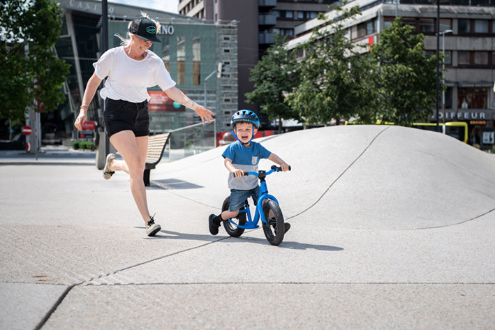 Kind auf blauem Laufrad und erwachsene Begleitperson im Freien. Urbanes Umfeld, geeignet für Kinderaktivitäten.