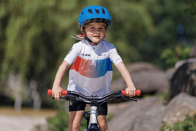 Kind auf Cube Kinderfahrrad mit blauem Helm, trägt Cube Trikot. Outdoor-Fahrradtour. Zielgruppe: Kinder.