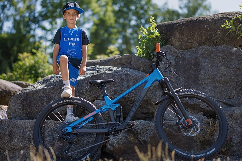 Junge in blauem Cube-Radtrikot neben einem blauen Cube-Kinderfahrrad auf Felsen im Freien.