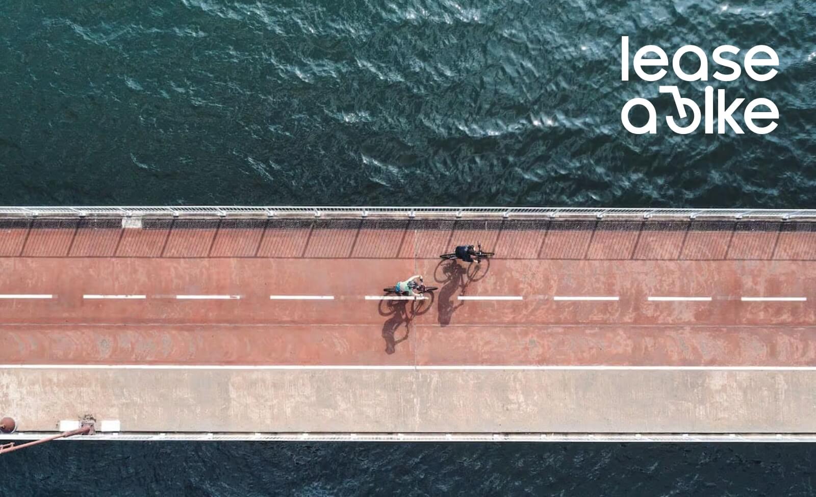 Zwei Personen fahren auf einer Brücke Fahrrad, Wasser im Hintergrund, mit "lease a bike" Schriftzug oben rechts.