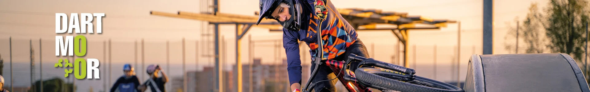 BMX-Fahrer im Helm springt in Skatepark bei Sonnenuntergang; Dartmoor-Logo sichtbar.