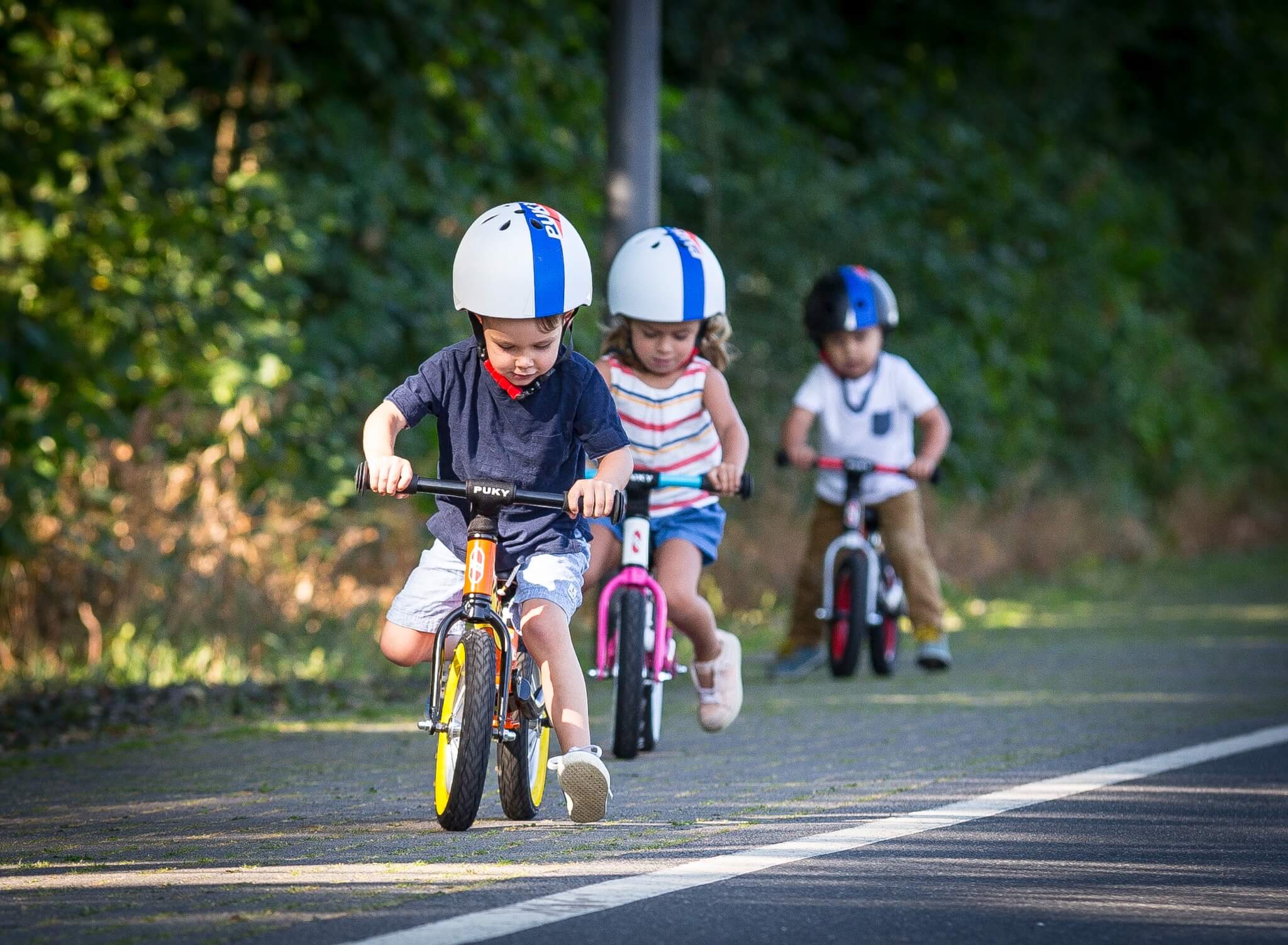 Kinder fahren auf Puky-Laufrädern mit Helmen auf einer Straße im Freien.