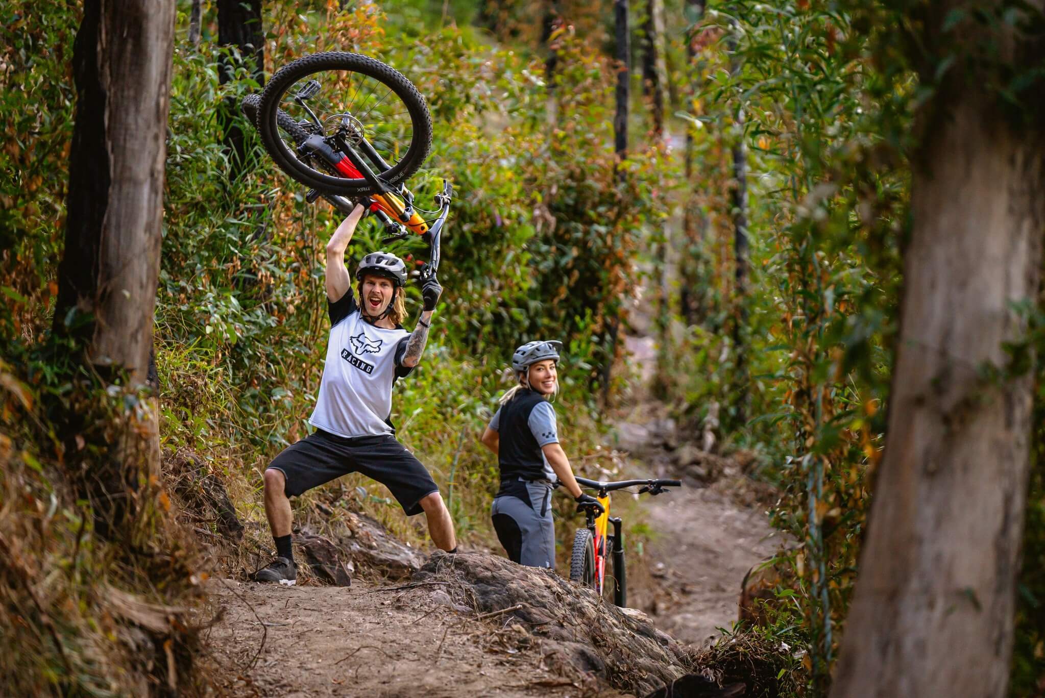Zwei Mountainbiker mit Trek Top Fuel im Wald, einer hebt begeistert sein Fahrrad hoch. Abenteuer im Gelände.