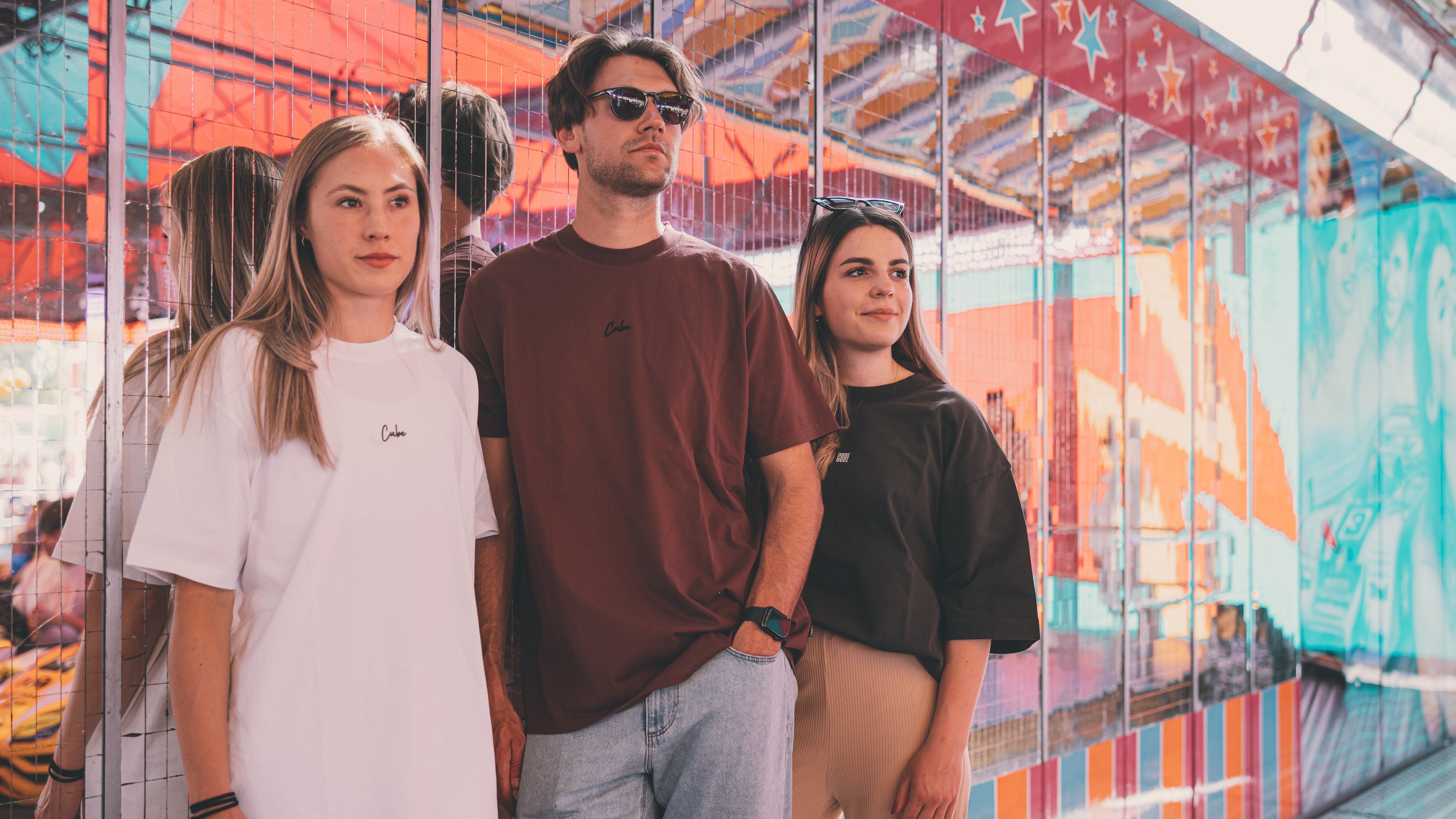 Drei Personen in Cube-Bekleidung T-Shirts stehen vor einem bunt beleuchteten Hintergrund bei einem Jahrmarkt.