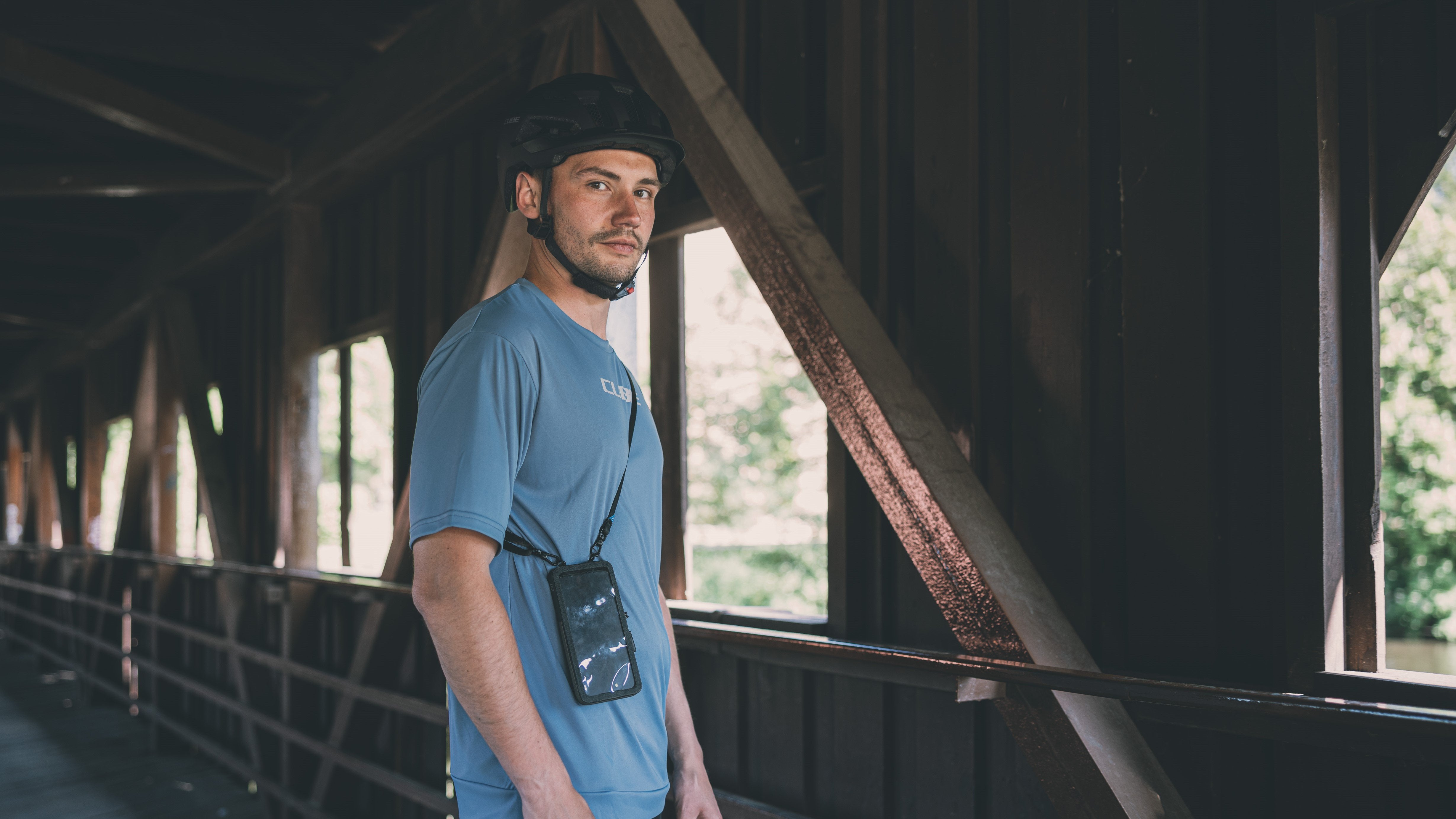 Mann mit Fahrradhelm und Cube-Logo auf blauem Shirt, steht mit Umhängetasche in überdachter Holzbrücke.