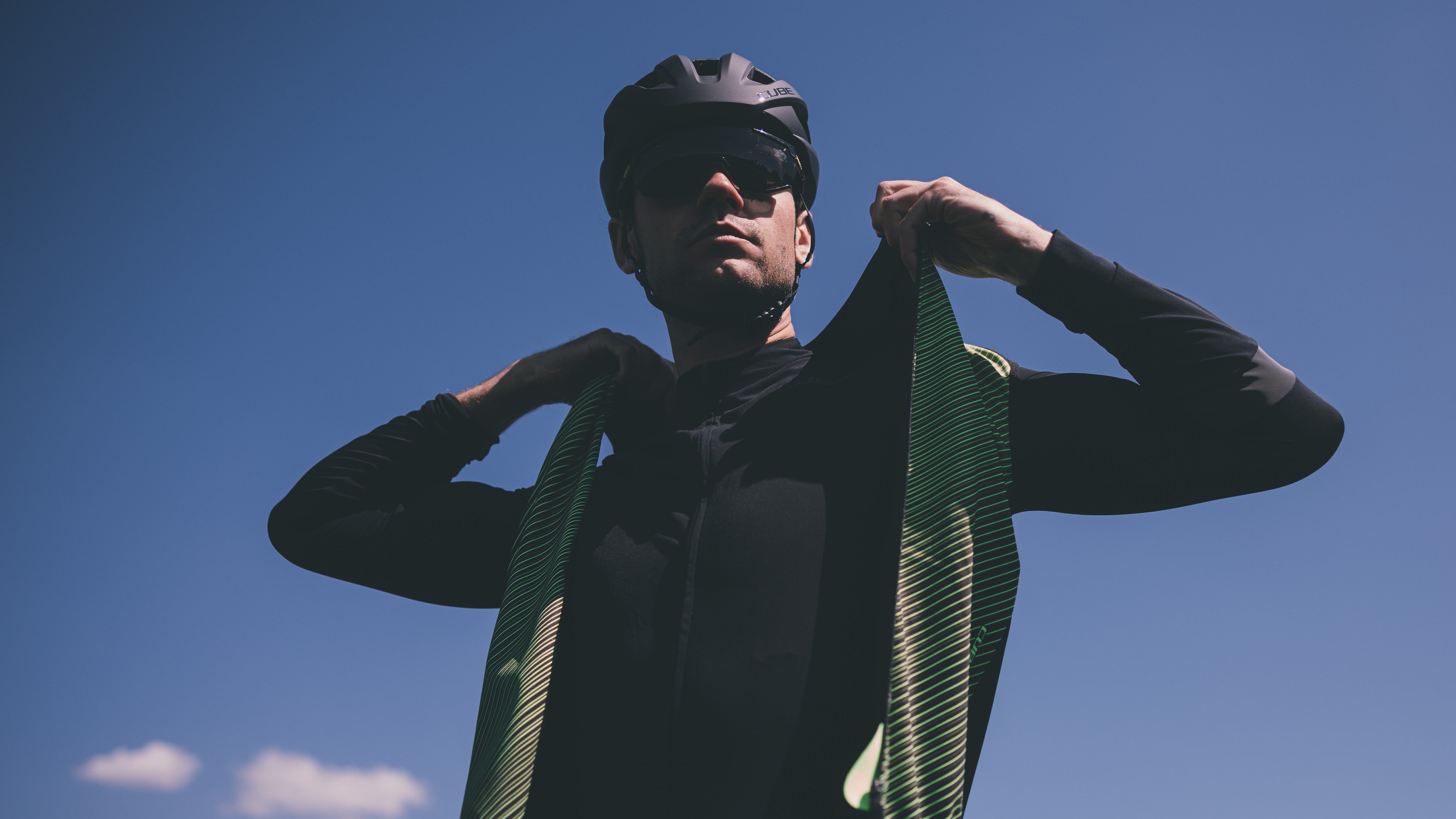 Radfahrer mit Cube-Helm und Sonnenbrille zieht schwarze Jacke vor blauem Himmel an.