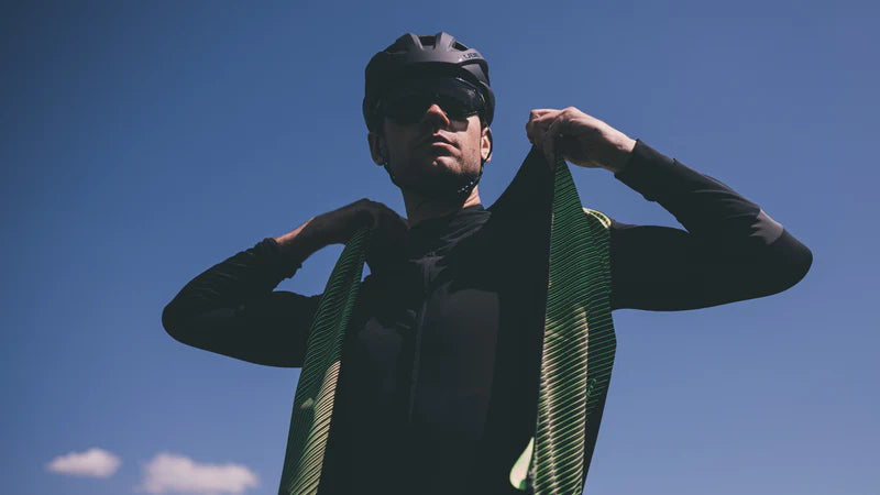 Person beim Anziehen einer sportlichen, gestreiften Fahrradweste unter blauem Himmel, trägt Helm und Sonnenbrille.